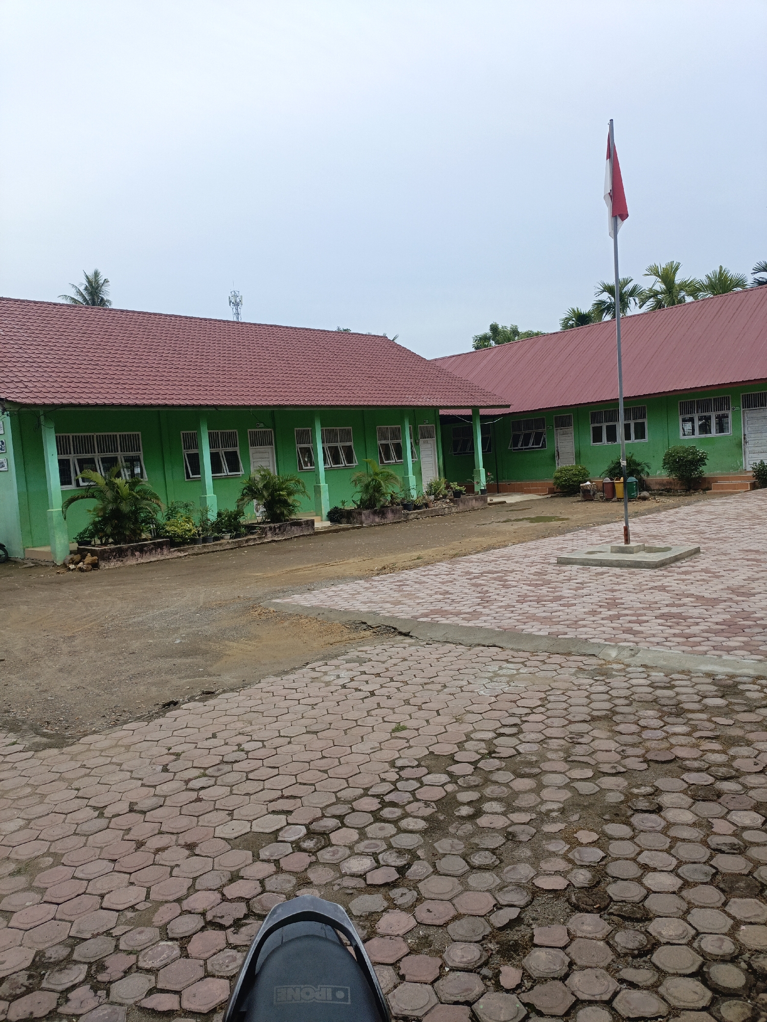 The image depicts the exterior of a school building in Lhokseumawe, Indonesia. The school has a distinctive green facade and a red tiled roof. In the foreground, a paved area with hexagonal interlocking bricks is visible, suggesting a courtyard or walkway. A flagpole with the Indonesian flag flying at half-mast stands prominently in the paved area. Several trees, including palm trees, are scattered around the school grounds, and a communication tower can be seen in the distance. The sky is overcast, indicating a cloudy day. The perspective suggests the photo was taken from a seated position, possibly on a motorcycle, as the back of a black seat with visible text is in the lower part of the frame. The overall scene conveys a typical school environment in a tropical region.
