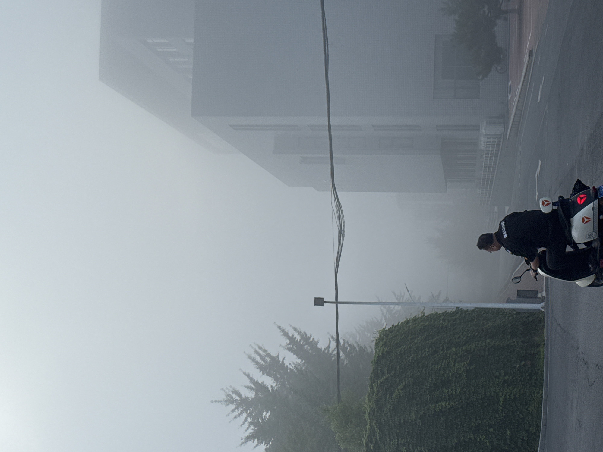 In Zhongtun, China, a man wearing a black uniform with white text, possibly identifying him as a security guard, rides a white scooter on a foggy day. He is moving from left to right across the frame. In the background, a large building with a brick facade is partially obscured by the fog, as are several trees. A dense green hedge lines the left side of the road. Electrical wires are strung across the scene, supported by a pole. The road surface is asphalt.