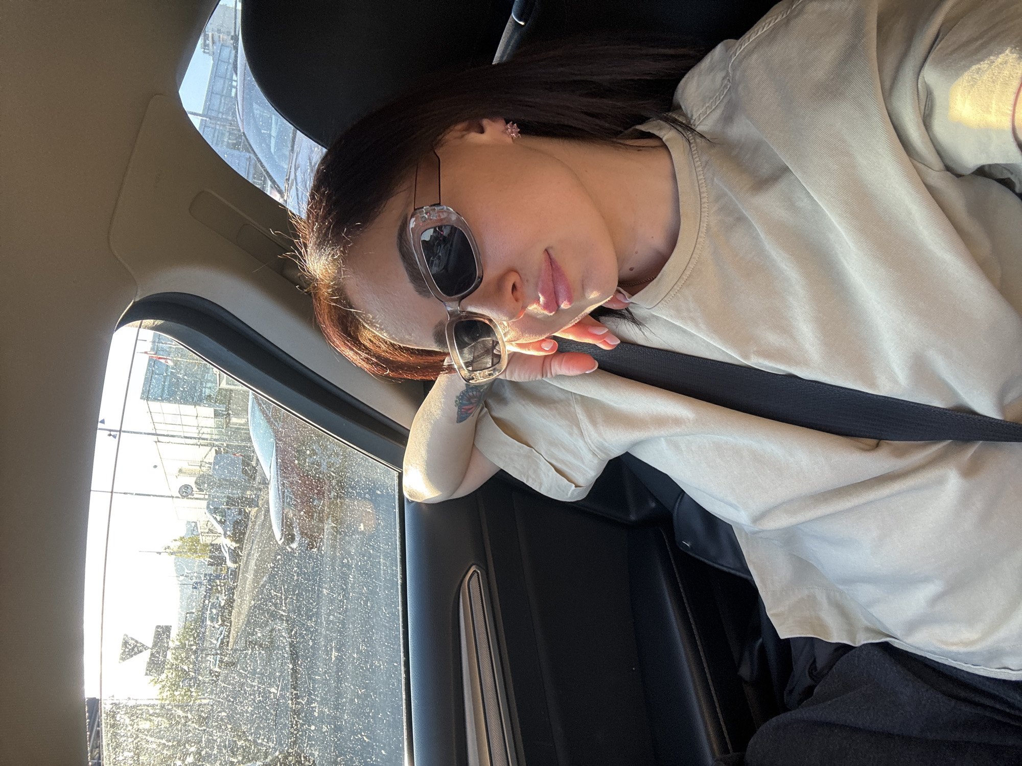 A young woman is shown reclining in the passenger seat of a car, viewed from the chest up with her head tilted. She has long dark hair and wears clear-framed sunglasses with dark lenses, her lips slightly parted in a relaxed expression. She wears a light-colored, possibly beige or light olive, short-sleeved T-shirt, a subtle gold chain around her neck, and a small pink, flower-shaped stud earring in her left ear. Her right forearm, adorned with a colorful tattoo (possibly a butterfly), rests on the dark interior door panel. A black seatbelt is visible across her chest.
Bright, natural sunlight strongly illuminates her face and arm, suggesting a sunny daytime. The car interior appears dark. Through the car window, which is speckled with dirt or water spots, an outdoor scene is visible. It depicts what looks like a parking lot or an urban area with distant buildings, other vehicles, and some scattered greenery under the bright sky. No discernible text is visible within the image.