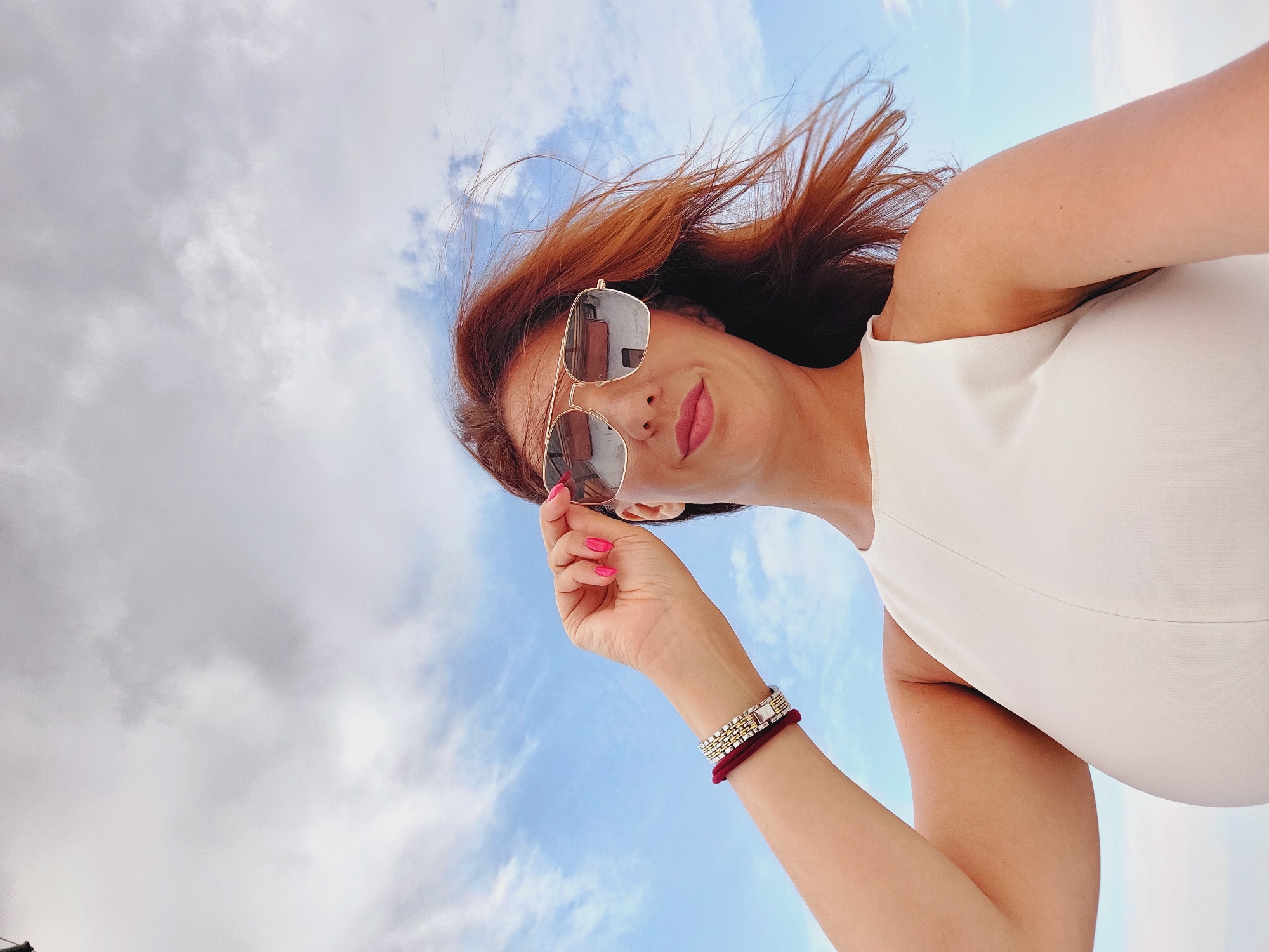 The image is a low-angle, selfie-style photograph of a woman with auburn hair and fair skin, taken outdoors under a partly cloudy sky, possibly in Sozopol, Bulgaria. She is wearing large, mirrored aviator sunglasses and a sleeveless white top. Her right hand is raised, adjusting her sunglasses, and her fingernails are painted bright pink. She has two bracelets on her left wrist: one appears to be a silver-toned link bracelet, and the other is a dark red band. Her hair is windswept, with strands blowing across her face. The background is dominated by a blue sky with scattered white and gray clouds. The overall impression is one of a bright, breezy day.