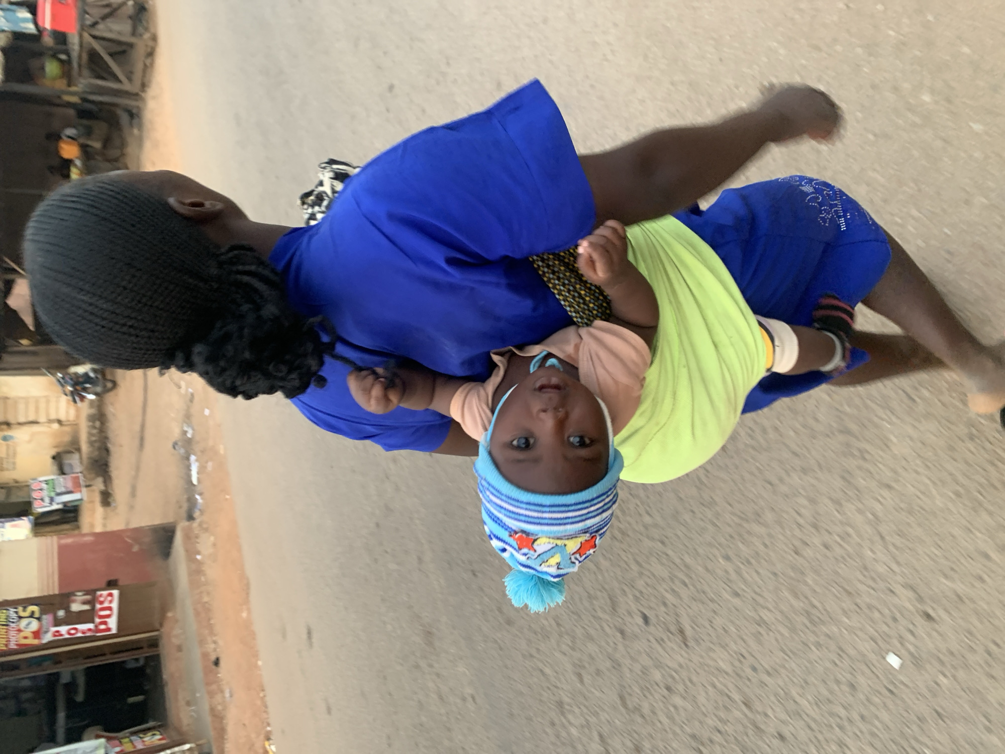 This image, taken in Jos, Nigeria, features an adult carrying an infant in an outdoor street setting. The adult, with dark skin and dark braided hair, is wearing a bright royal blue top and matching blue shorts or trousers. A section of fabric with a dark, possibly polka-dotted or patterned, design is visible between the adult's top and the baby carrier.
The infant, also with dark skin, is carried in a vibrant neon green fabric sling or carrier, facing forward. The baby is dressed in a light peach-colored top and a distinctive light blue and white striped beanie adorned with multicolored patterns and a fluffy blue pom-pom on top. A light blue pacifier is in the baby's mouth, and its wide, attentive eyes are looking directly at the camera.
The background reveals a typical street scene with a light gray-brown paved or unpaved road surface. On the left side, several buildings, likely shops or stalls, are visible. Signage on these buildings includes clear instances of "POS" (Point of Sale) and partially legible text suggesting "PRINTING PHOTOCOPY" services, indicating local businesses. A vehicle, possibly a motorcycle, is faintly discernible in the distance. The scene is illuminated by natural light, indicating daytime. The photograph captures a candid moment of daily life, with the baby's direct gaze being a notable detail.