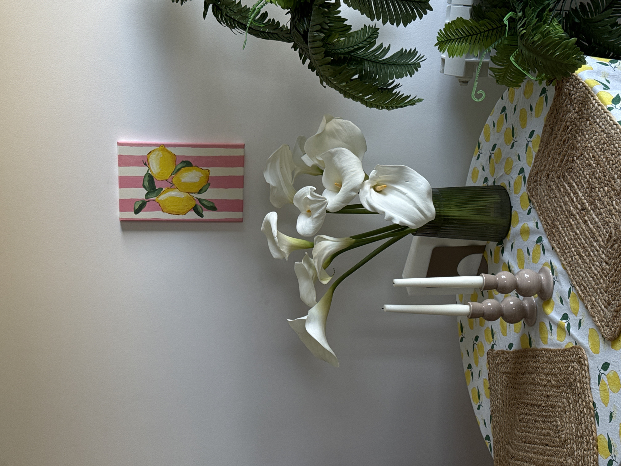 The image shows a still life arrangement. A lemon-themed painting hangs on a light-colored wall. Next to it is a vase of calla lilies. Two candle holders sit on a lemon-patterned tablecloth, partially covered by a woven placemat. Green foliage hangs overhead. The location is not discernible from the image.