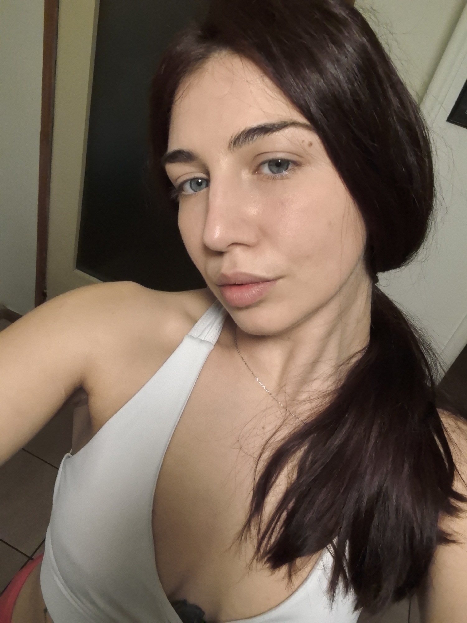 A close-up, indoor shot depicts a young woman with light skin, striking blue eyes, and dark brown hair pulled back in a ponytail. She is wearing a white halter top and a delicate gold necklace. Her facial expression is neutral. The background includes a dark-colored door on the left and a white wall on the right. The location is impossible to determine from the given visual information. No information relates to the city of Stara Zagora, Bulgaria.
