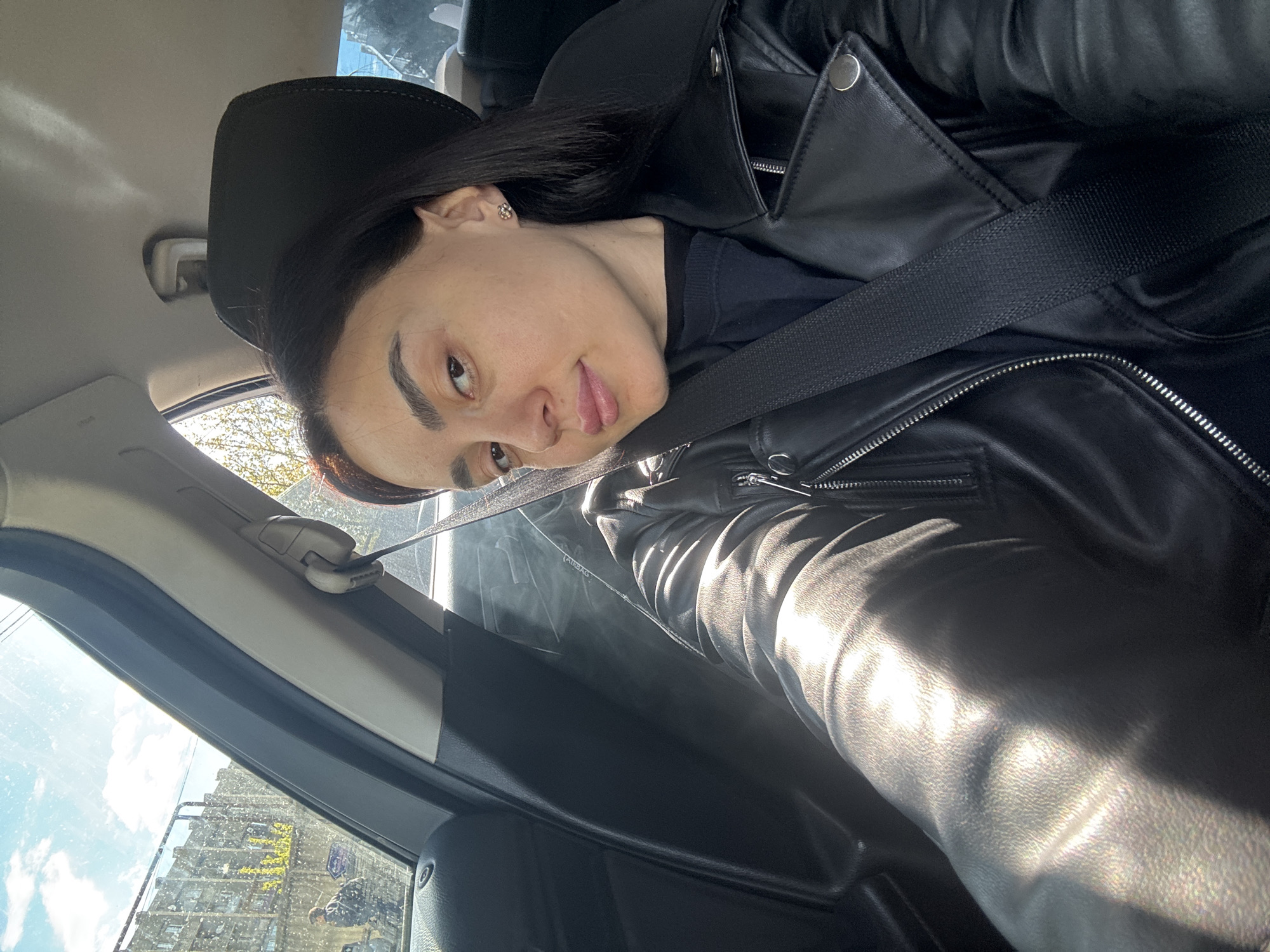 A woman is seated in the driver's seat of a car, wearing a black leather jacket and a seatbelt. She is looking directly at the camera. Outside the car window, a building is visible. Based on building style and surroundings, the location is likely Tbilisi, Georgia.