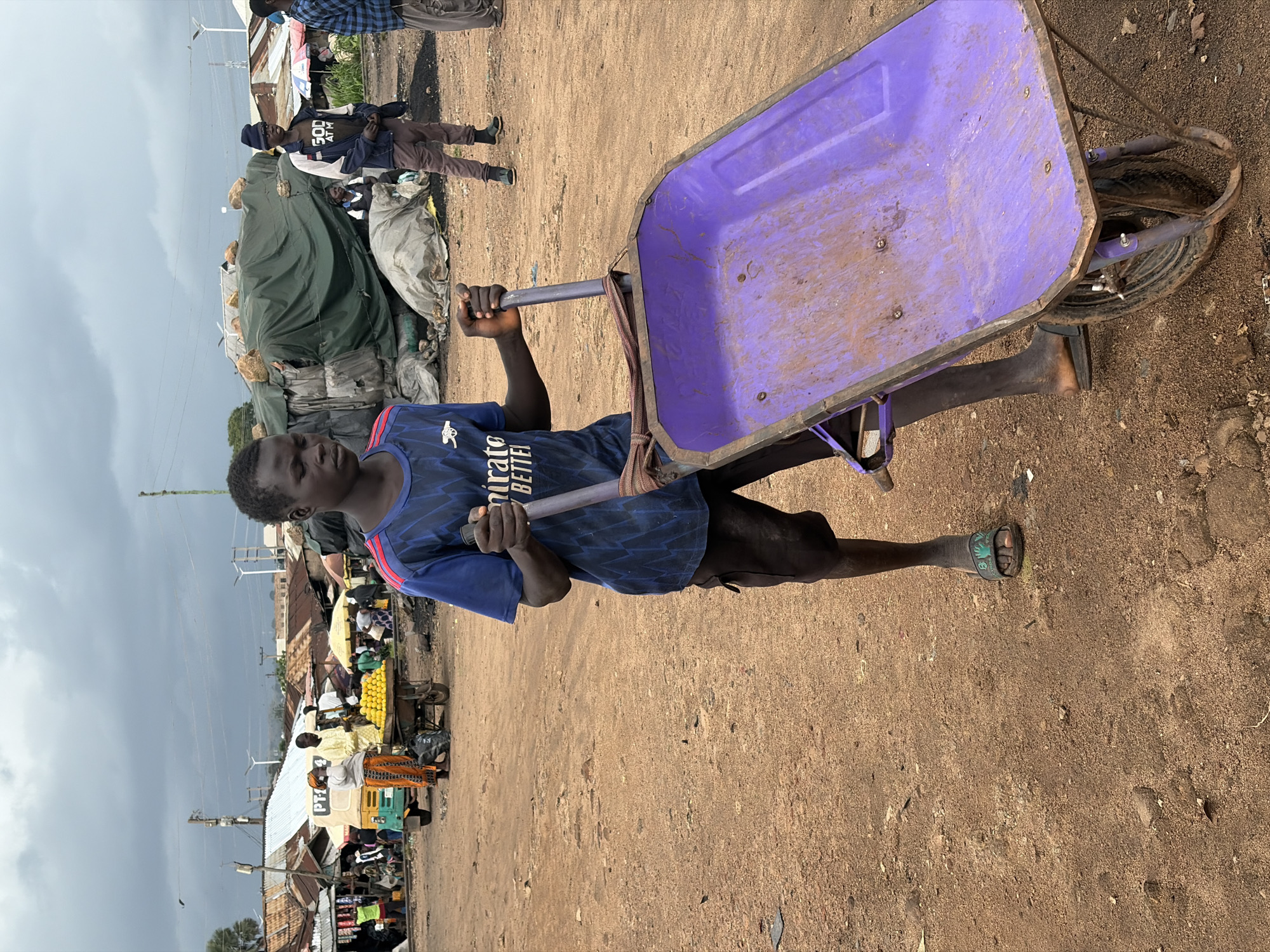 In Bukuru, Nigeria, a young boy, appearing to be of teenage years, stands on a dirt road holding a purple wheelbarrow. He wears a blue t-shirt with an Arsenal logo and red trim, and dark shorts. His footwear consists of black slides. The wheelbarrow is loaded with a tarp, suggesting it is used for transport or storage. In the background, a bustling market scene unfolds with various stalls, makeshift structures covered by tarps, and other individuals going about their business. A three-wheeled vehicle, a tuk-tuk, is visible, as are piles of what appear to be fruits, possibly mangoes, for sale. The sky is overcast, indicating a cloudy day. The overall environment suggests a vibrant, active marketplace in a semi-urban or rural setting within Nigeria.