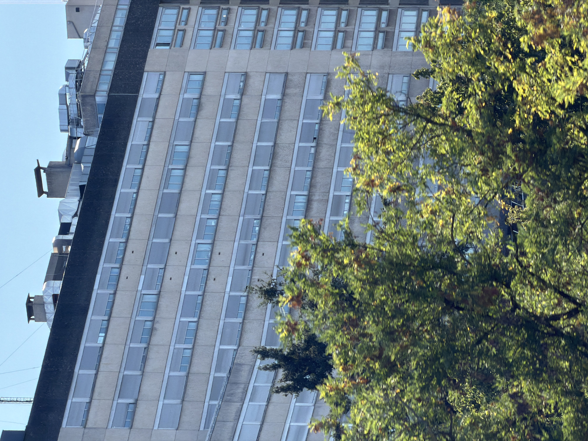 The image shows the facade of a multi-story building in Bron, France. The building appears to be an older, institutional structure, possibly a hospital or office building, constructed from concrete panels. Numerous windows, covered with horizontal blinds, are arranged in rows across the facade. The rooftop features visible HVAC units and ventilation systems, as well as chimneys. In the foreground, lush green trees with some autumnal foliage obscure parts of the building, suggesting the photo was taken from a street or park adjacent to the structure. The sky is clear and blue. There are no people visible in the image.