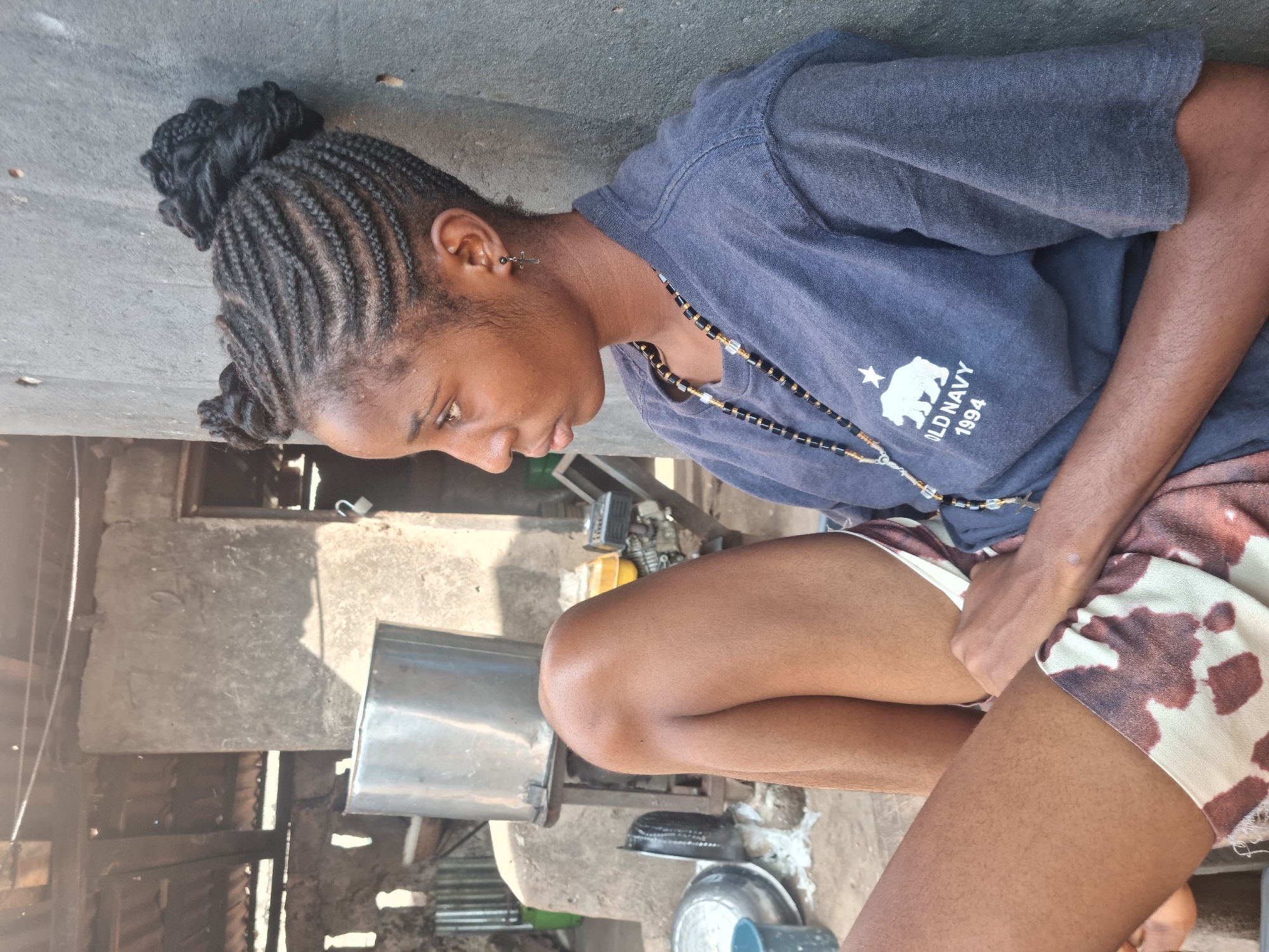 This media file captures a young Black woman in an outdoor setting in Onitsha, Nigeria, during bright daylight hours. She is shown from the waist up, seated or squatting, looking downwards with a neutral, possibly contemplative, expression.
The woman has black hair styled in neat cornrows that gather into a bun at the back of her head. She is wearing a dark blue t-shirt adorned with a white graphic featuring a polar bear, a star, and the text "OLD NAVY 1994." Accessories include a silver-colored cross-shaped earring in her left ear and a beaded necklace composed of black, gold, and clear beads. Her lower attire consists of shorts with a distinctive brown and white cow-print pattern.
The background reveals a functional, somewhat rustic environment. There are concrete surfaces and structures made of what appears to be concrete blocks, wood, and corrugated metal. A large metallic silver container, possibly a pot or industrial vessel, is visible next to her. Further back, parts of machinery, resembling an engine or generator, are present. A blue bowl or basin is partially visible on the ground. The strong sunlight suggests it is midday or early afternoon. The woman's hands are resting in her lap, and no specific activity is clearly depicted.