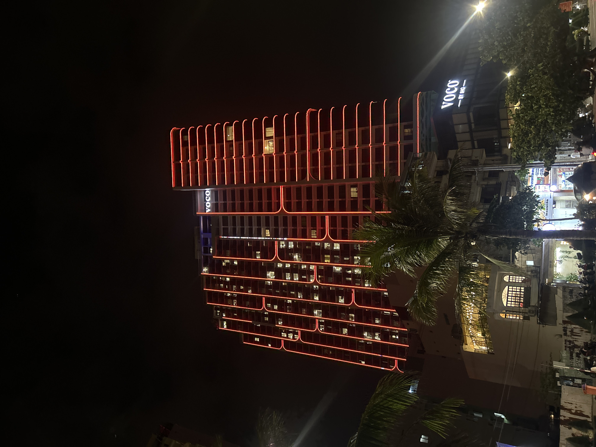 The original image, taken at night, captures a striking modern high-rise building, identified as "VOCO by IHG" by its prominent illuminated sign near the top. The building is adorned with distinctive red-orange LED or neon light strips that outline its architectural structure, creating a grid-like pattern with U-shaped elements on each floor. Numerous windows throughout the building are lit from within, suggesting occupancy.
In the foreground and to the right, large palm trees partially obscure the base of the main building and other surrounding structures. Beyond these trees, glimpses of a lively urban environment are visible, including other illuminated buildings, bright streetlights, and what appear to be commercial establishments. The sky above is a deep, unlit black, emphasizing the artificial glow of the city. No specific human activity or event is discernible; the focus is on the illuminated architecture and the nighttime ambiance of the location. The scene depicts a vibrant resort or city district at night, consistent with its location in Bắc Nha Trang, Vietnam.