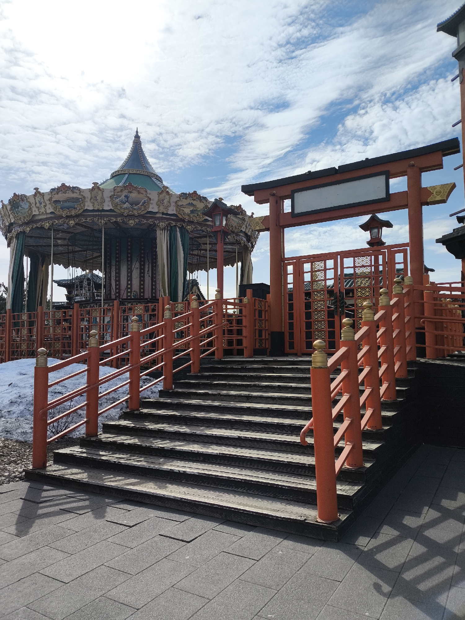 The image shows a carousel and a traditional Japanese torii gate in a park-like setting. The carousel is in the background, featuring a classic design with ornate decorations and a green and brown roof. A partially visible snow patch suggests a recent winter season.
In the foreground, a set of wooden stairs leads up to the torii gate, which is painted in a vibrant orange color. The gate has a blank white sign board on top. A wooden fence with matching orange posts and railings lines the steps. The ground is covered with gray paving stones. The sky above is bright blue with scattered white clouds. The location is Izmailovo Kremlin in Moscow, Russia.