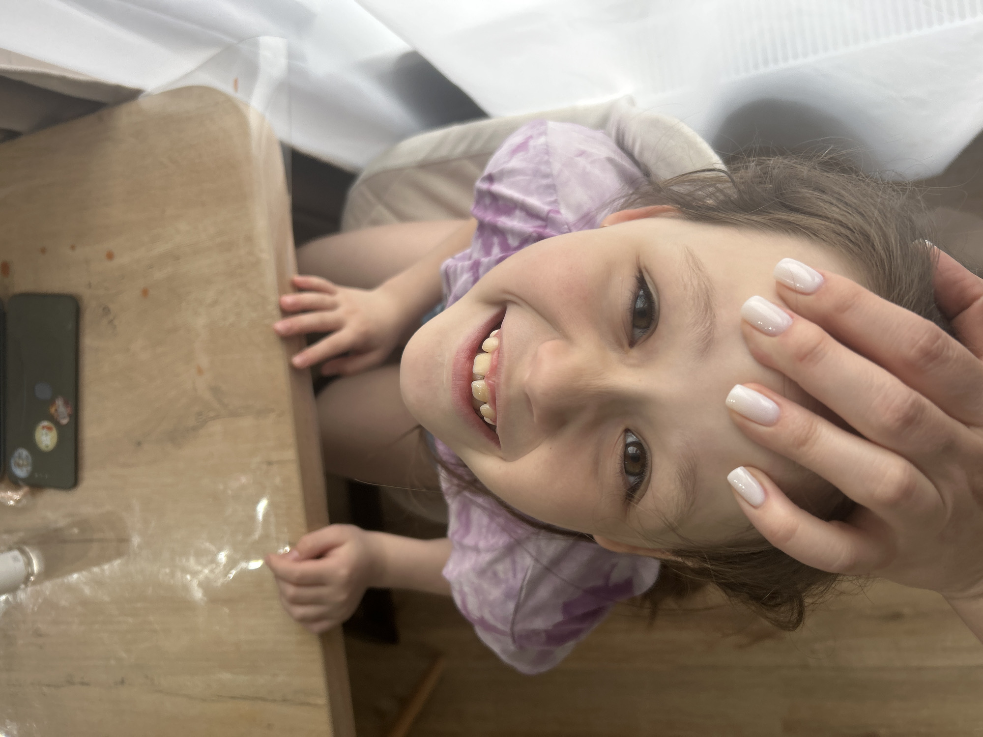 A young girl is smiling and looking upwards at the camera. Her head is partially obscured by a hand with white manicured nails, indicating an adult likely taking the picture. She is wearing a tie-dye purple shirt and is seated in what appears to be a white chair. In the background, there is a wooden table covered with a clear plastic sheet. A black mobile phone can be seen on the table. There is also what looks like white curtains overhead. The scene appears to be inside a home. There is no information in the image to verify the location is Chaikovsky, Russia.