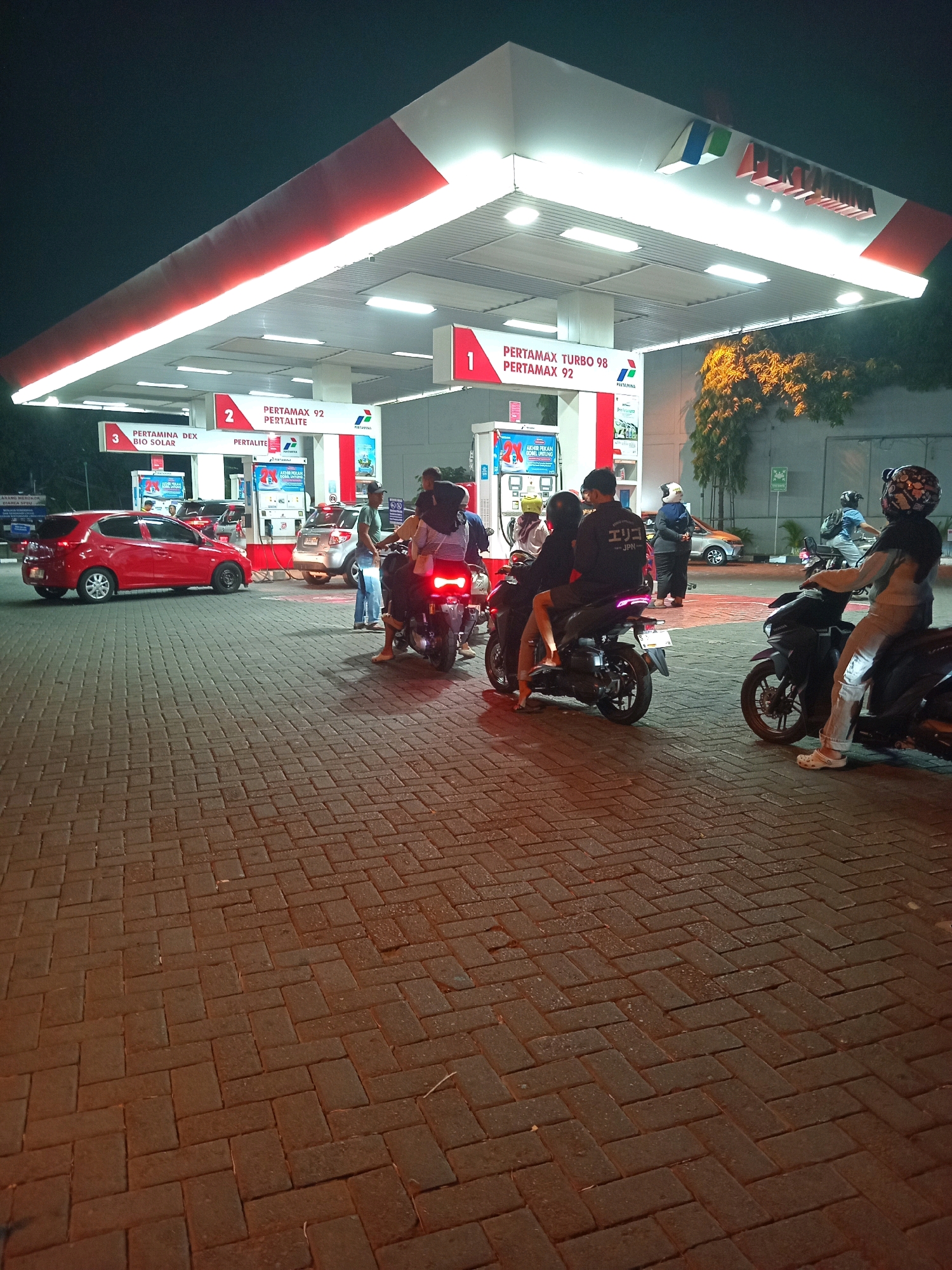 The image depicts a Pertamina gas station at night, likely in Karangturi, Indonesia, as indicated by signs. The scene is illuminated by the bright lights of the gas station canopy, casting a warm glow on the brick-paved ground. Several people are present, most of them on motorcycles.
In the foreground, two motorcycles are parked side-by-side. The one on the left has two people seated on it, facing towards the gas pumps. The rider is wearing a dark jacket with Japanese text on the back, and the passenger has a backpack. The motorcycle on the right has a single rider wearing a helmet and light-colored pants, also facing the pumps.
Behind these motorcycles, a red car is parked at a gas pump, with its doors open and seemingly being refueled. Another car, silver in color, is also at a pump. Several other individuals are visible around the gas pumps, some appearing to be attending to the vehicles or interacting with each other.
The gas station features prominent signage with the Pertamina logo and information about different fuel types like "Pertamax Turbo 98," "Pertamax 92," "Pertamina Dex," and "Bio Solar." A woman in a blue headscarf and dark clothing stands near a parked car in the background, observing the scene. The overall environment suggests a typical, active gas station in an urban or suburban setting at night.