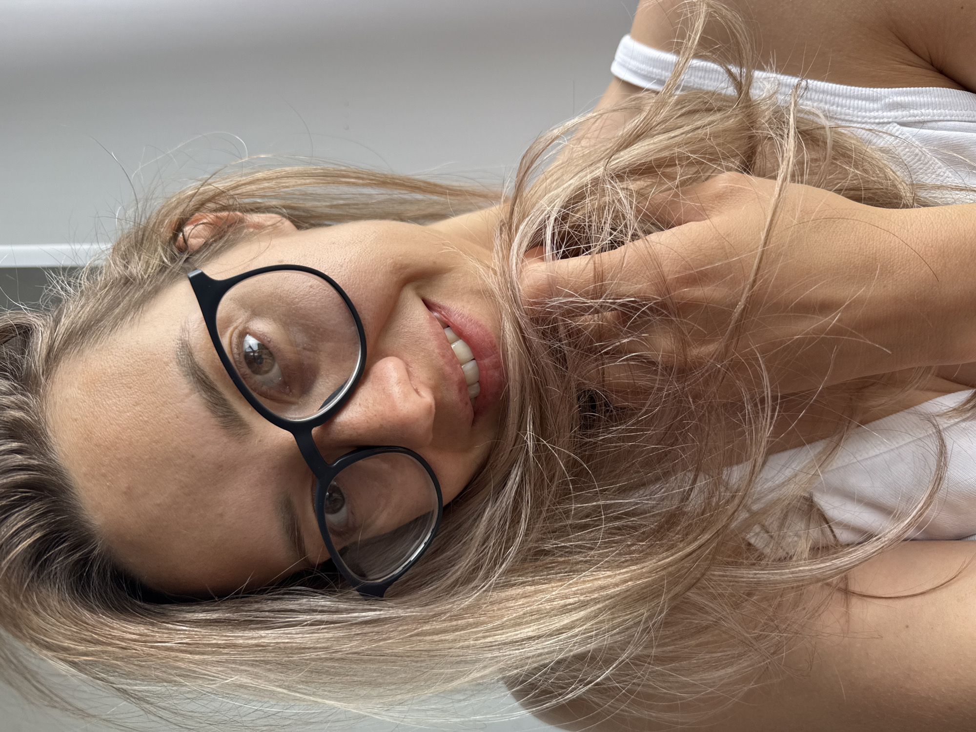 A woman with blonde hair, wearing glasses and a white tank top, is shown from the chest up, smiling with her hand in her hair. The background is a plain, light-colored wall. There is no discernible information in the image that could indicate the location in Chaikovsky, Russia.