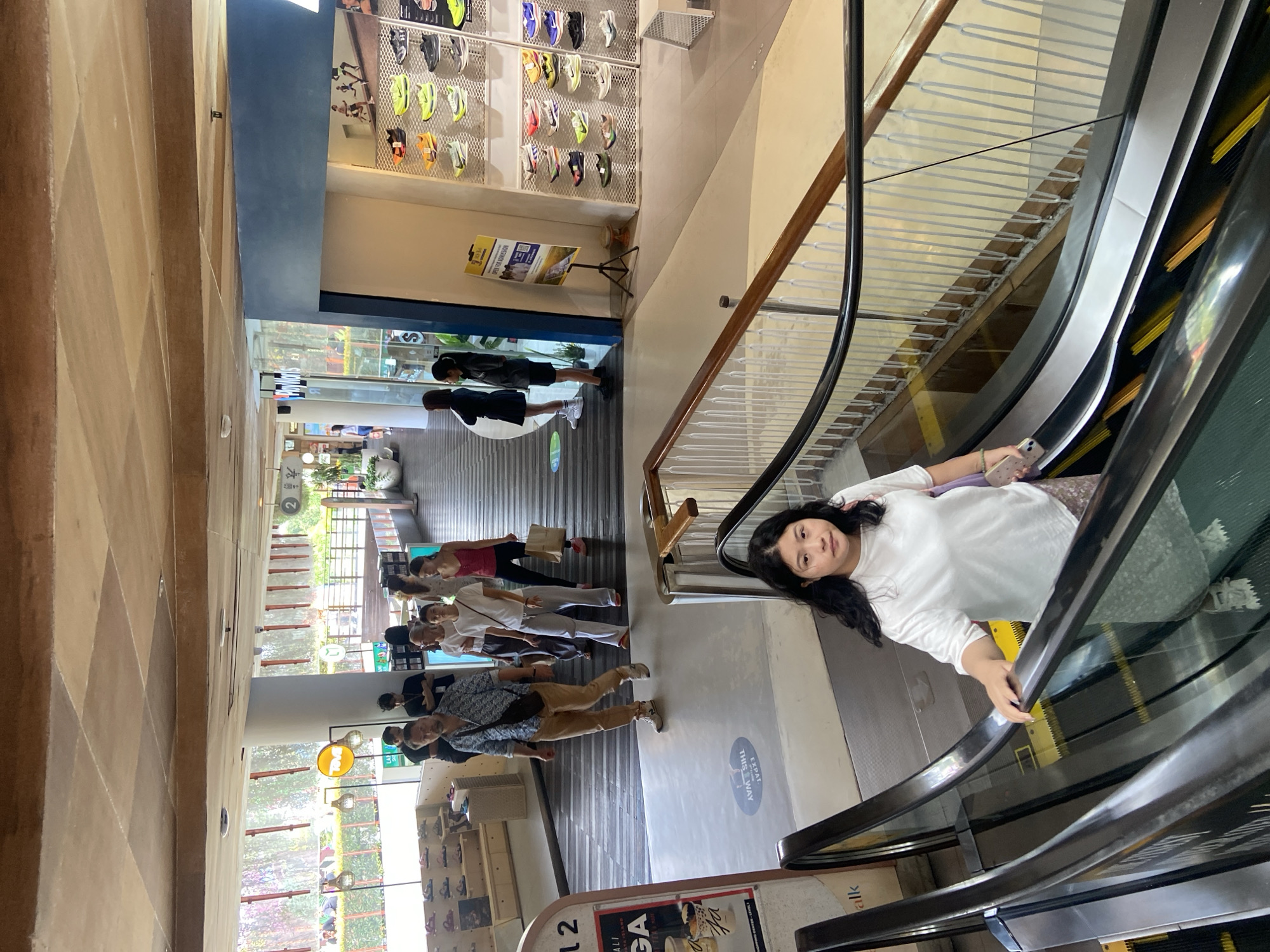 The image shows a young woman with long, dark, curly hair riding an escalator in what appears to be a shopping mall in Kuta, Indonesia. She is wearing a white t-shirt and a patterned skirt, and is holding a smartphone in her left hand. She is looking towards the camera with a neutral expression. The escalator is black with yellow treads. In the background, other shoppers are visible, and a store selling athletic shoes is displayed on the right. The shopping mall has a modern design with exposed wooden beams on the ceiling and large windows. The lighting is bright, suggesting it is daytime.