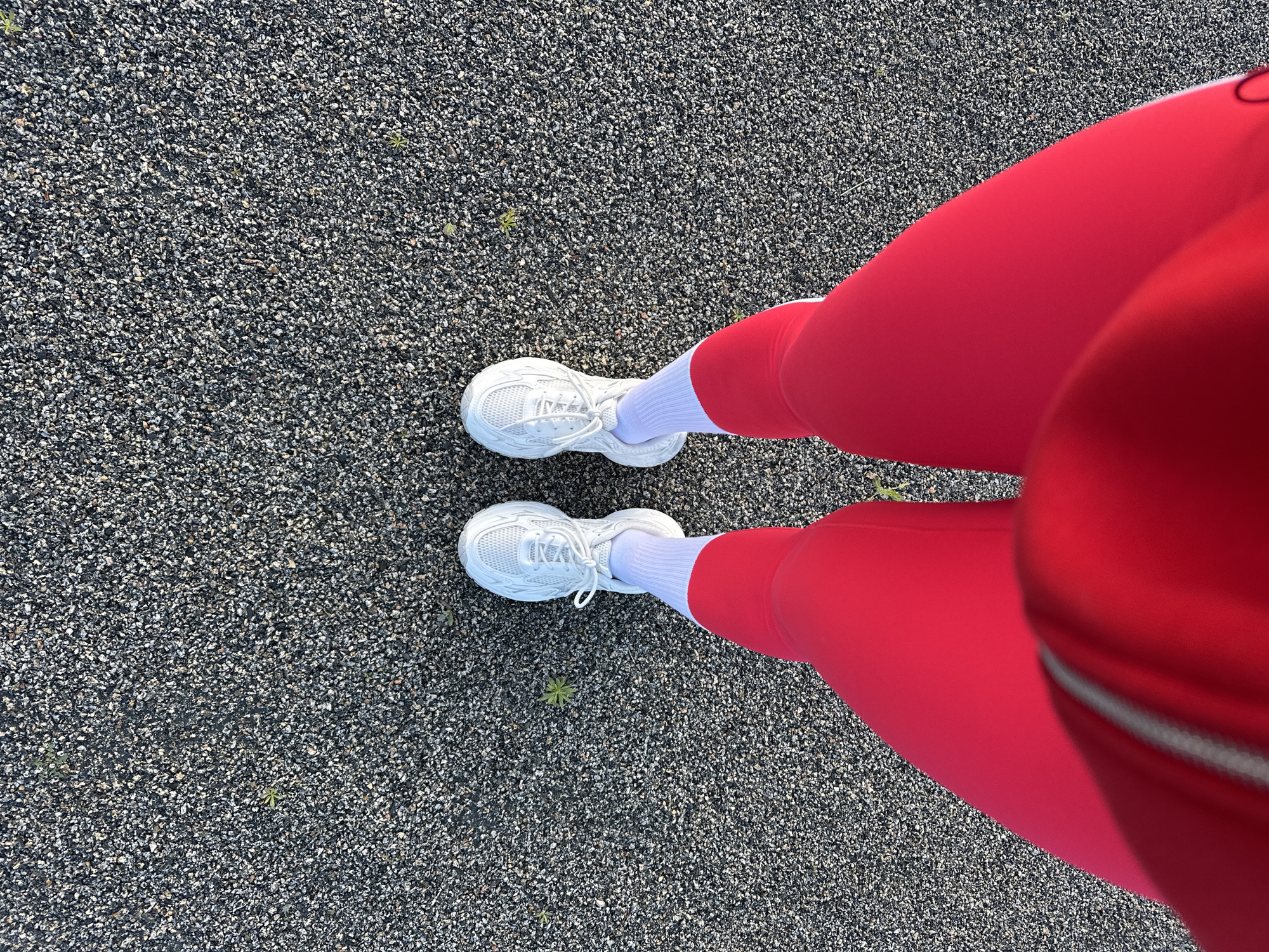 The image presents a top-down view of a person's lower body, specifically their feet and lower legs, standing on a dark, textured ground surface that appears to be asphalt or fine gravel. Scattered across the ground are small, sparse green plants. The person is wearing white athletic sneakers with visible laces and white crew-length socks that extend above the ankle. Bright red leggings or pants cover the remainder of the visible legs, with a subtle lighter-colored seam or zipper detail visible on the garment in the foreground. This scene is located in the city of Yuzhnouralsk, Russia.