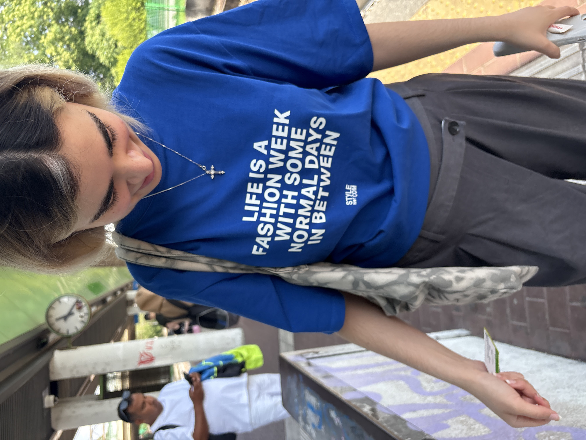 A young woman stands outdoors, wearing a royal blue t-shirt with the white text "LIFE IS A FASHION WEEK WITH SOME NORMAL DAYS IN BETWEEN," along with a small logo at the bottom right reading "STYLE NOT COM." She also wears dark gray pants with a button closure and a silver chain necklace with a cross pendant. She has fair skin and her hair is dyed with a blonde fringe. A patterned shoulder bag is slung across her body. She is holding a small white object in her right hand.
In the background, partially visible, is a platform with a clock on a pole. A man wearing a white shirt and sunglasses is standing to the left. Based on the clock and the presence of the metro, the location appears to be a metro station in Milan, Italy.