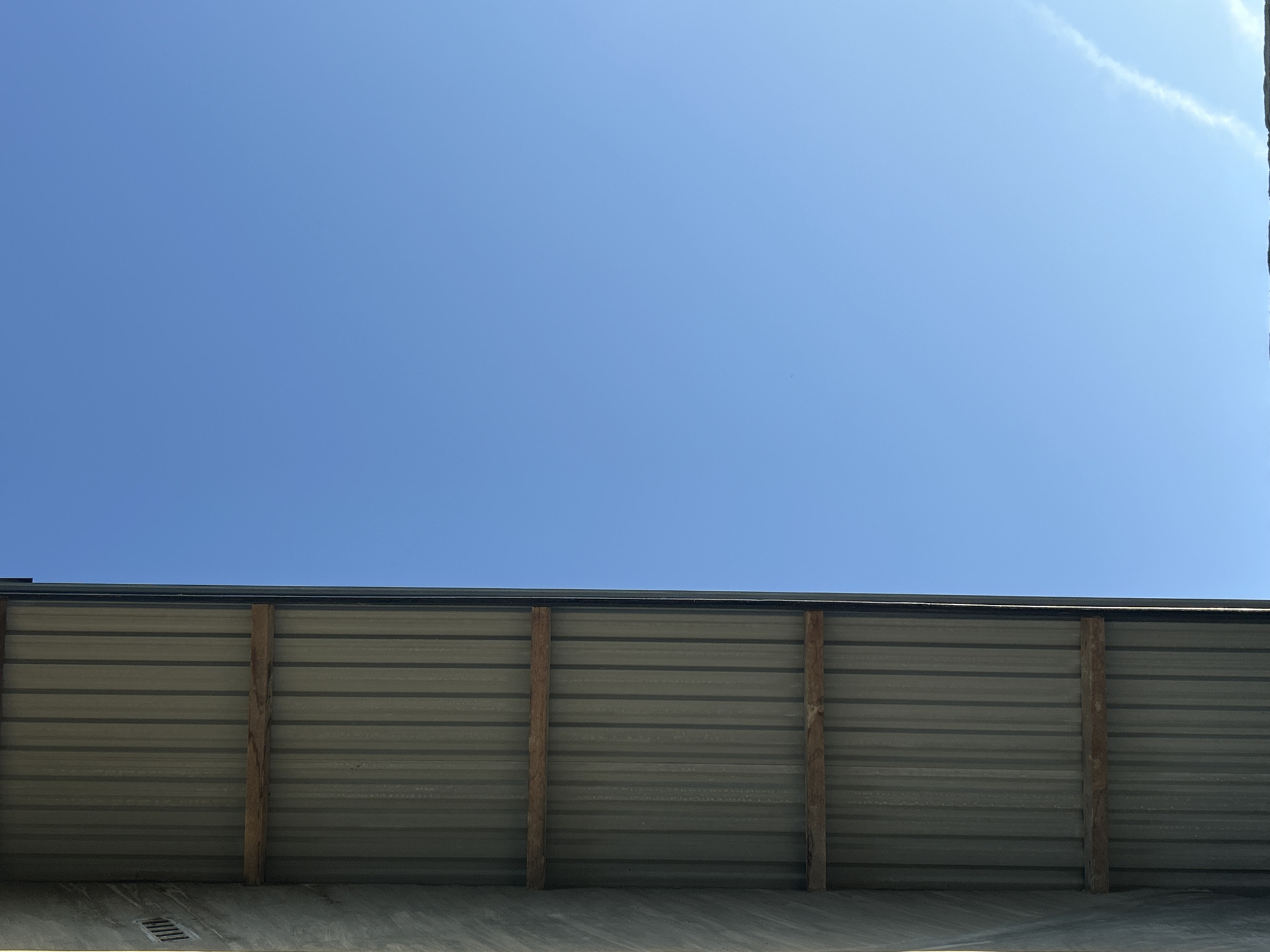 The image depicts a low-angle view looking upwards at the corner of a building, likely in an outdoor setting in Lhokseumawe, Indonesia. The building's exterior wall is constructed of corrugated metal panels, vertically supported by wooden beams. The lower portion of the wall rests on a concrete or paved surface. The upper section of the view is dominated by a clear blue sky with a faint wisp of a cloud in the upper right corner. There are no visible people, subjects, or interactions in the frame. The focus is on the architectural details and the open sky above.