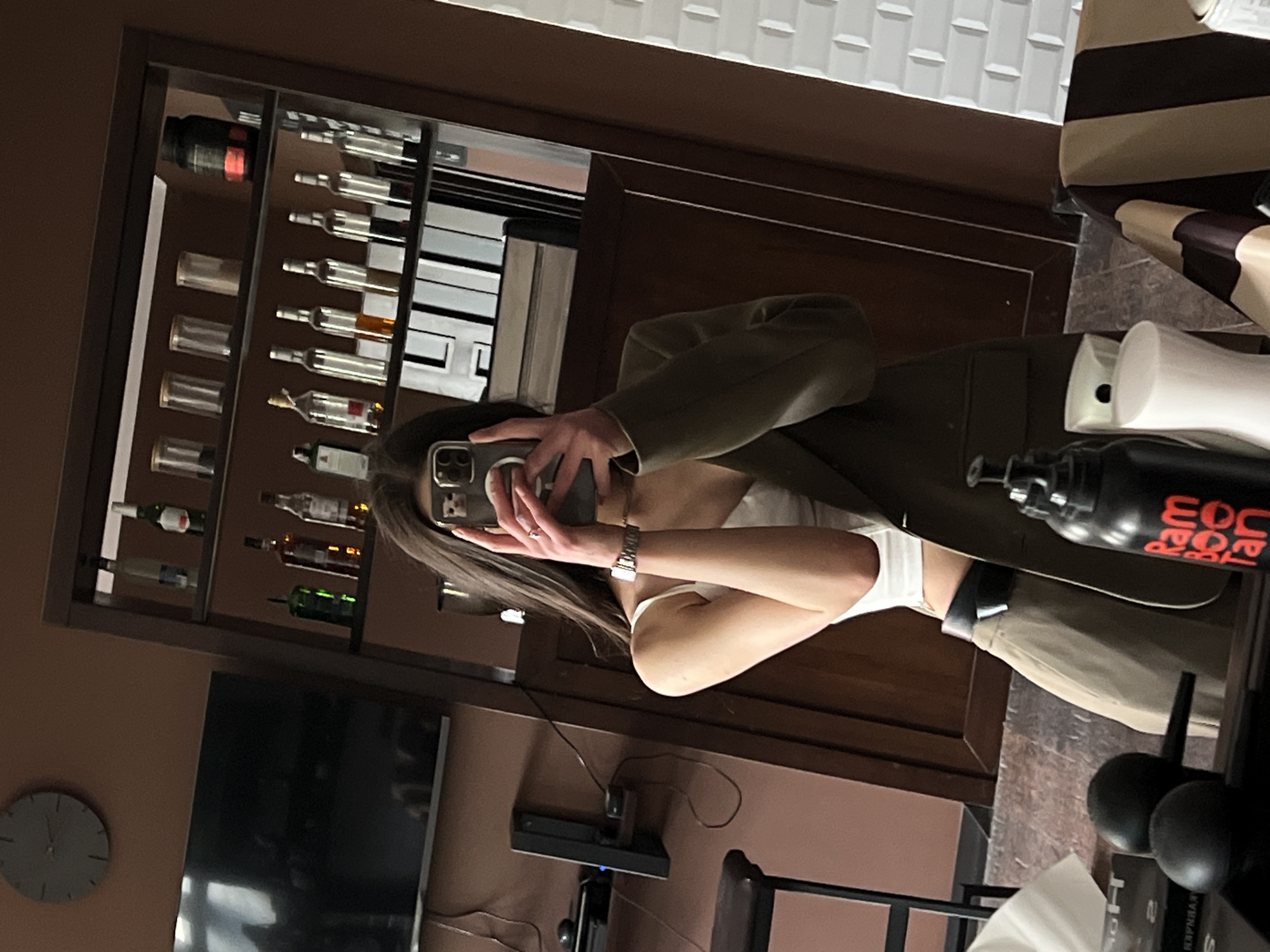 The photograph, taken in Ternopil, Ukraine, depicts a woman holding a smartphone and capturing a selfie in a mirror. The woman is wearing a white top and a light olive green jacket. Her face is partially obscured by the phone she is holding up. Behind her is a wall with a display case stocked with various bottles of what appears to be alcoholic beverages, along with a flat-screen television and a clock. In front of the woman, near the edge of the mirror's reflection, are various salon-related items such as hair products and a hair dryer. The environment suggests the photograph was taken inside a salon or a similar business establishment. The floor has a dark wood appearance.