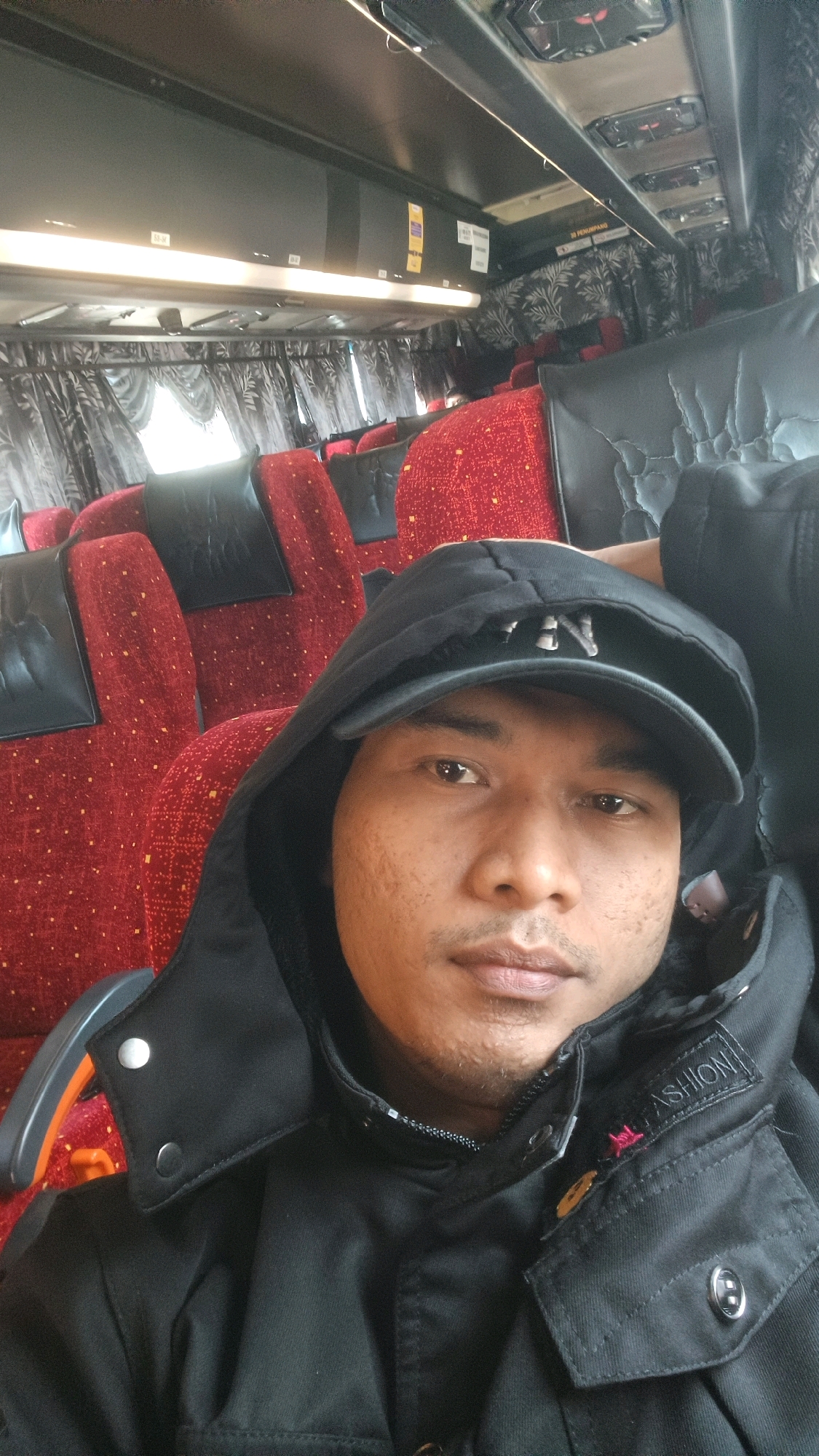 The image is a selfie taken inside a bus, likely in Permatang Pauh, Malaysia. The bus interior features rows of red and black seats with ornate patterns on the red upholstery and black leather headrests. The ceiling of the bus is black and houses overhead compartments and lighting. The individual in the foreground is wearing a black hooded jacket and a black baseball cap. Their face is visible, showing brown eyes, a light complexion with some blemishes, and a mustache and stubble. The person is looking directly at the camera. The overall lighting suggests it is daytime.