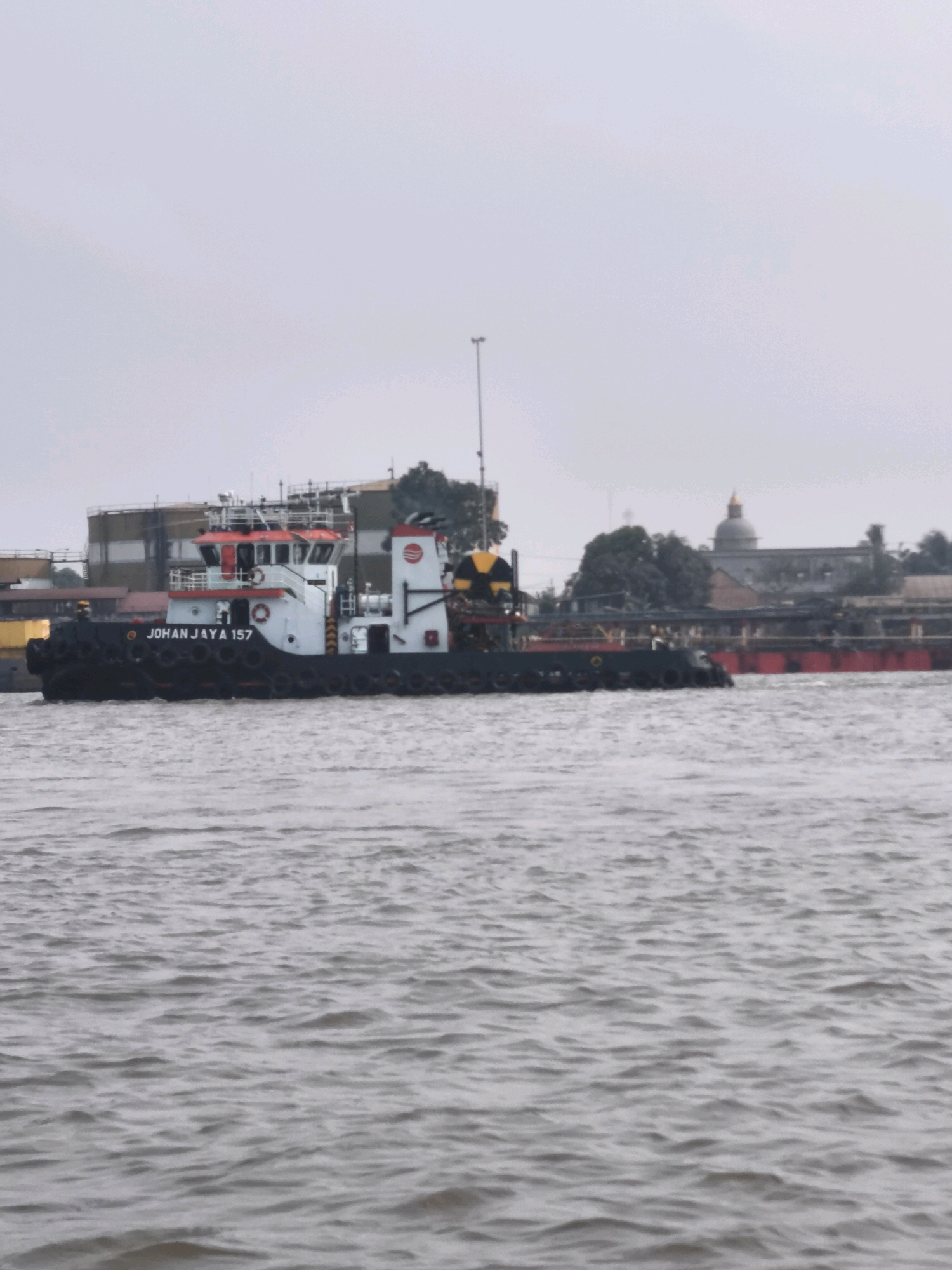 A tugboat named "JOHAN JAYA 157" is pictured in the Musi River in Palembang, Indonesia. The tugboat is white with red accents and a black hull, with large black tires around its sides. It is towing a barge filled with what appears to be cargo. In the background, industrial structures and a building with a dome are visible along the riverbank. The sky is overcast.
