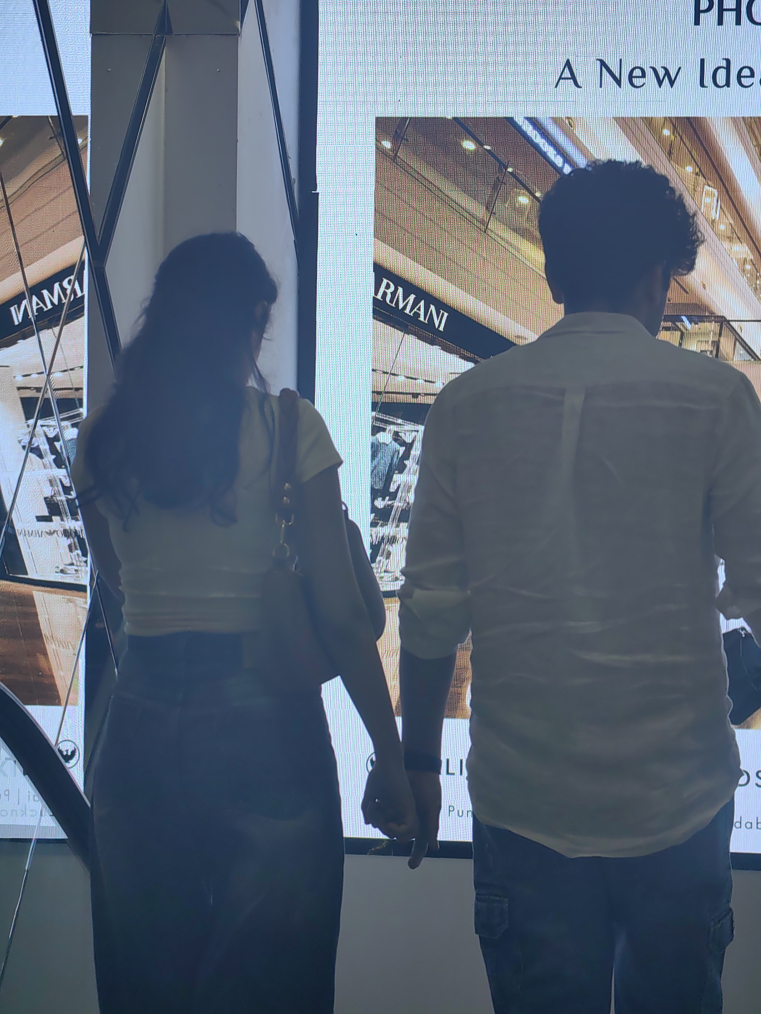 A man and a woman are seen from behind, holding hands, in front of a large digital screen displaying an advertisement. The man is wearing a light-colored, long-sleeved shirt and dark pants. The woman is wearing a light-colored top and dark pants. The digital screen shows text and images, including the word "ARMANI". The overall scene suggests an indoor location, possibly a shopping mall or a public space. The reflection on the screen suggests the presence of other elements in the environment. The image appears to be taken at the Jio World Drive mall in Mumbai, India, based on the ARMANI advertisement and the overall aesthetic commonly found in such locations.