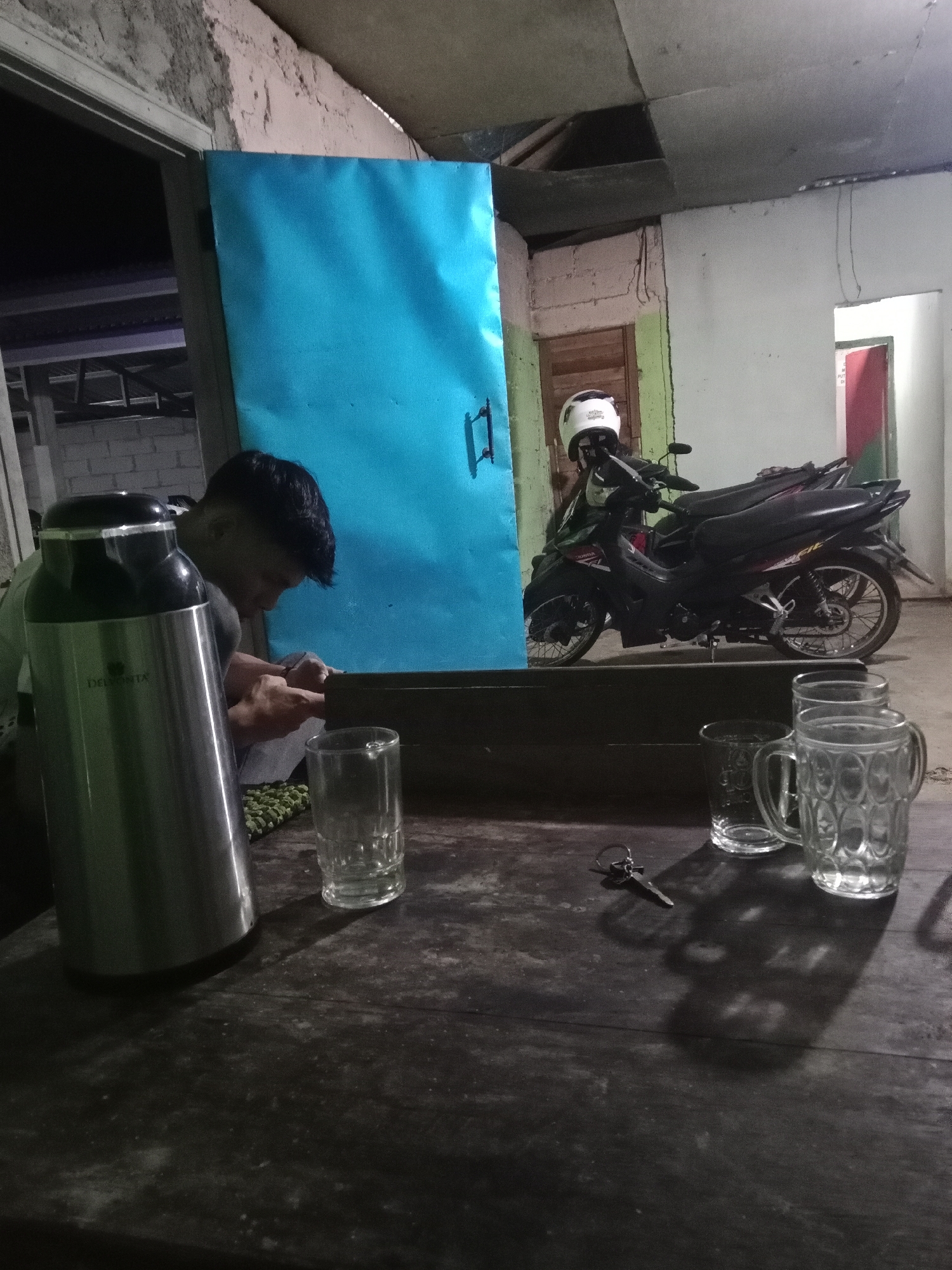 The image depicts an indoor scene, likely a simple roadside establishment or shelter, in Leksono, Indonesia. In the foreground, a person with dark hair, wearing a light-colored shirt, is seated at a wooden table, appearing to be engaged in an activity involving their hands. In front of them is a large silver thermos with a black lid, and a pile of what appears to be green beans. To the right of the person, on the table, are two empty clear glasses, one of which is a mug with a handle. A single key lies on the table near the glasses.
The background reveals a blue door or panel, a portion of a white brick wall, and a motorcycle parked indoors. The motorcycle is black with silver accents and has a white helmet resting on its handlebars. The environment suggests a rustic or utilitarian setting, with visible imperfections in the walls and ceiling. The lighting is dim, creating shadows that contribute to the overall atmosphere of the scene. The presence of the motorcycle and the open doorway suggest a transition point between indoor and outdoor spaces.