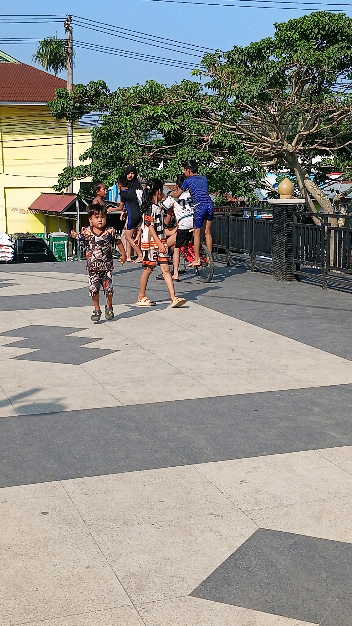 In Sungai Tarab, Indonesia, several children are gathered on a paved area with a distinctive geometric pattern. A young boy in the foreground is mid-jump, wearing a patterned t-shirt and shorts. Behind him, a group of children are interacting. One child is sitting on a red bicycle, with another child standing behind them, possibly assisting. Other children in the background are observing or participating in the activity. A tree with dense green foliage is visible behind the group, and a yellow building with a red roof is present on the left side of the image. A black metal fence runs along the right side of the scene. The sky is clear and blue.