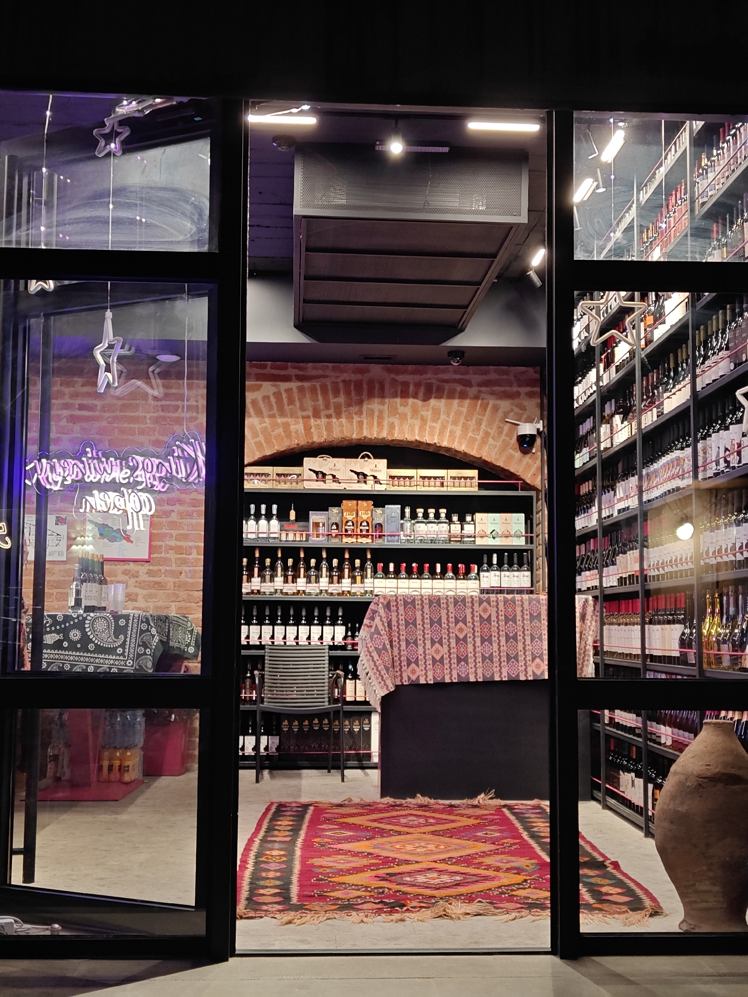 The image presents a view into a retail establishment, likely a wine and spirits shop, observed through its dark-framed glass storefront at night. The interior is brightly illuminated, contrasting with the unseen darkness outside.
On the left, through a large window pane, an exposed brick wall features a purple neon sign that appears to read "WINES, SPIRITS & BEER" with another word below, possibly "FOOD" or "GASTRONOMY." White, star-shaped decorative lights hang in the window. Below the neon sign, a table covered with a dark, bandana-patterned cloth holds several bottles and other items. A map is also visible on the brick wall.
The central section of the shop's interior showcases an exposed brick archway. Beneath it, shelves are filled with bottles of spirits and wines, and cardboard boxes, some labeled "Midiani," are stacked above. A security camera is mounted on the wall to the right of the arch. Overhead, an industrial-style ventilation unit is visible. In front of the shelves, a table or counter is draped with a colorful, patterned fabric, and a modern chair is positioned nearby. A large, intricate rug, rich in red, orange, and blue geometric patterns, covers the floor in this area.
The right side of the shop features tall, metal-framed shelves packed densely with an extensive collection of wine bottles, their labels and the ceiling lights reflecting on the glass. In the lower right foreground, a large, rustic clay pot, characteristic of a kvevri, stands on the floor, hinting at traditional Georgian winemaking.
No people are present in the scene, suggesting the shop might be closed or captured in a quiet moment. The combination of modern industrial elements, traditional decor, and the extensive display of bottles points to a specialized retail space, likely focused on beverages.