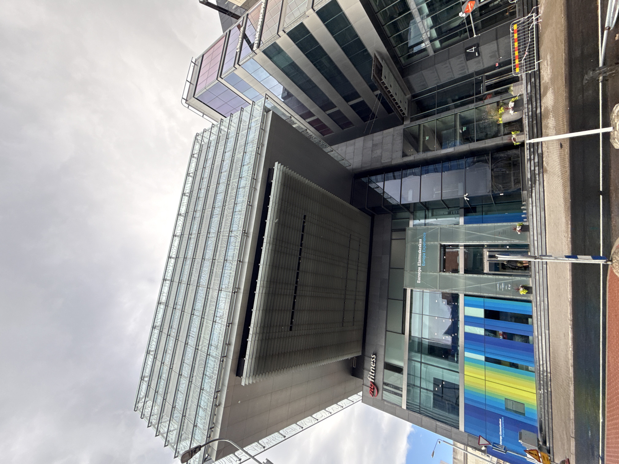 An upward-angle shot captures the modern architecture of the Europa Experience building in Tallinn, Estonia, under an overcast sky. The building features a striking design with overlapping geometric forms and extensive glass facades. A large, cantilevered section with a glass roof extends outwards, creating a dramatic overhang above the entrance. The lower part of the building displays a vibrant mural of blue and yellow stripes. A prominent red "myfitness" logo is visible on the side of the structure. Several people are seen near the entrance, one partially obscured by a glass door. Yellow caution signs and a temporary barrier are present on the sidewalk. The scene is typical of a commercial district in a European city, with the wet pavement suggesting recent rain.