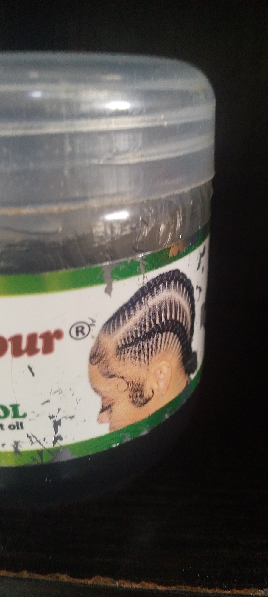 The image shows a close-up of a plastic container, likely hair product, with a label. The label features a graphic of a person's head with braided hair and text that is partially visible. The visible text includes "our" and "OL". The container is clear with a ribbed top. The background is dark and out of focus. There are no discernible elements that would indicate a specific location in Jalingo, Nigeria.