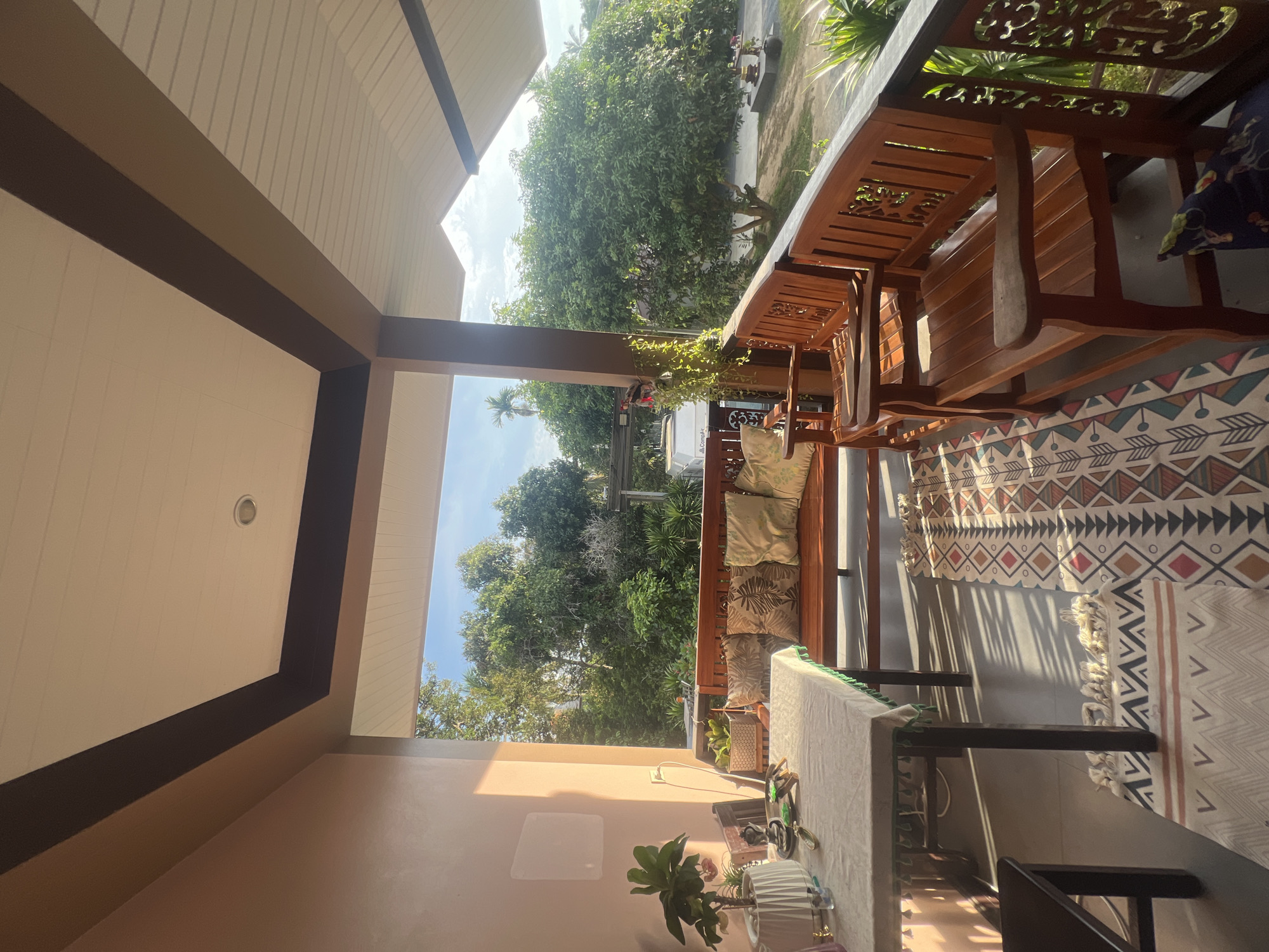 An outdoor covered patio in Ko Samui District, Thailand, features two wooden benches with cushions and a table covered by a patterned tablecloth. A rug with geometric patterns is laid on the floor. In the background, lush green trees and a bright sky are visible. Sunlight casts shadows across the scene.