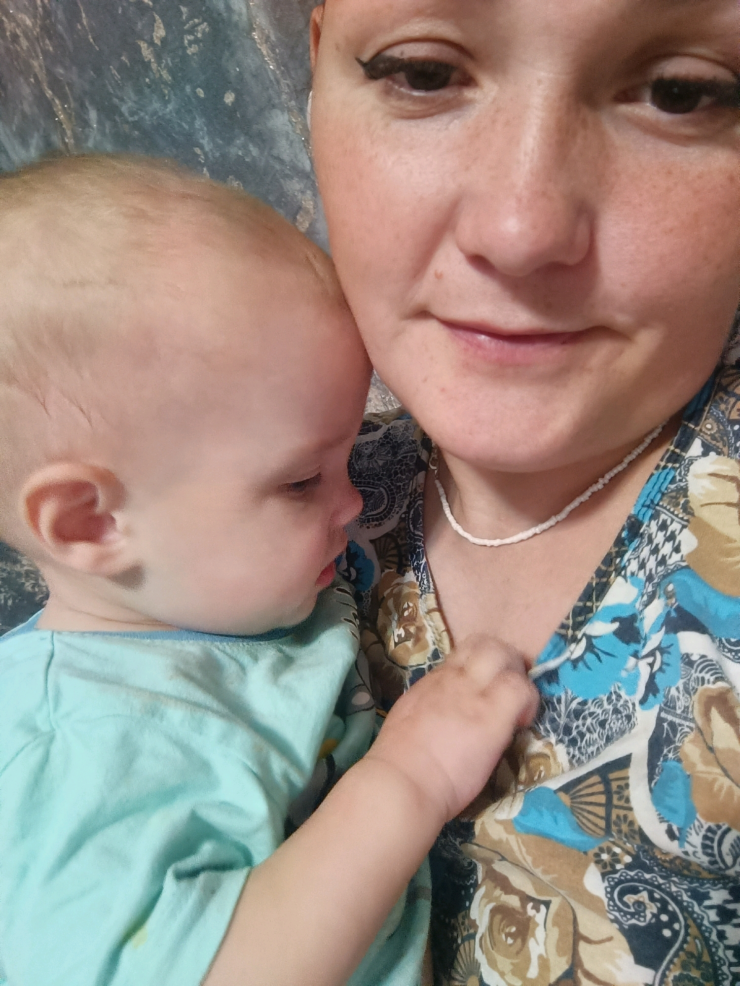 The image shows a close-up of a woman and a baby, likely a mother and child, in an indoor setting. The woman's face is in the foreground, showing her eyes, nose, and mouth. She has freckles and is wearing eye makeup. A thin white beaded necklace is visible around her neck, and she is wearing a patterned blouse with blue, brown, and white floral and paisley designs. The baby is positioned to the left of the woman, with its head resting on her shoulder or chest. The baby's face is mostly turned away from the camera, but its profile, ear, and part of its head are visible. The baby is wearing a light blue t-shirt. The background appears to be a wall with a textured, possibly marbled or painted surface in shades of blue and gray. The lighting is natural, casting soft shadows. There is no indication of the specific location within the city of Karsha, Russia, or any outdoor elements.