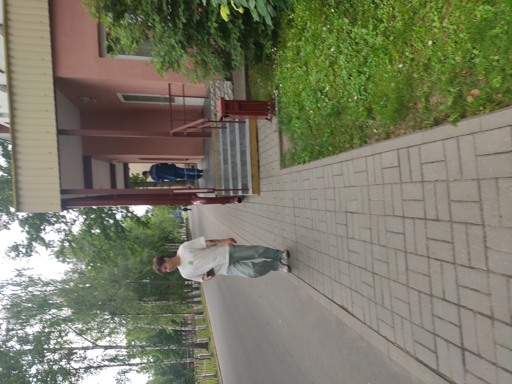 A young man with brown hair, wearing a white t-shirt and light blue baggy jeans, walks along a paved sidewalk in Navapolatsk, Belarus. He is holding a dark-colored smartphone in his right hand. Behind him, a person in a blue uniform walks up a set of outdoor stairs leading to a building with a salmon-pink facade. The building has a covered porch area with maroon support posts. To the right of the building entrance is a patch of green grass with some small white flowers and fallen leaves, and a dark red, metal trash receptacle. To the left of the sidewalk is a paved road bordered by a metal fence and trees with green foliage. The sky appears overcast.