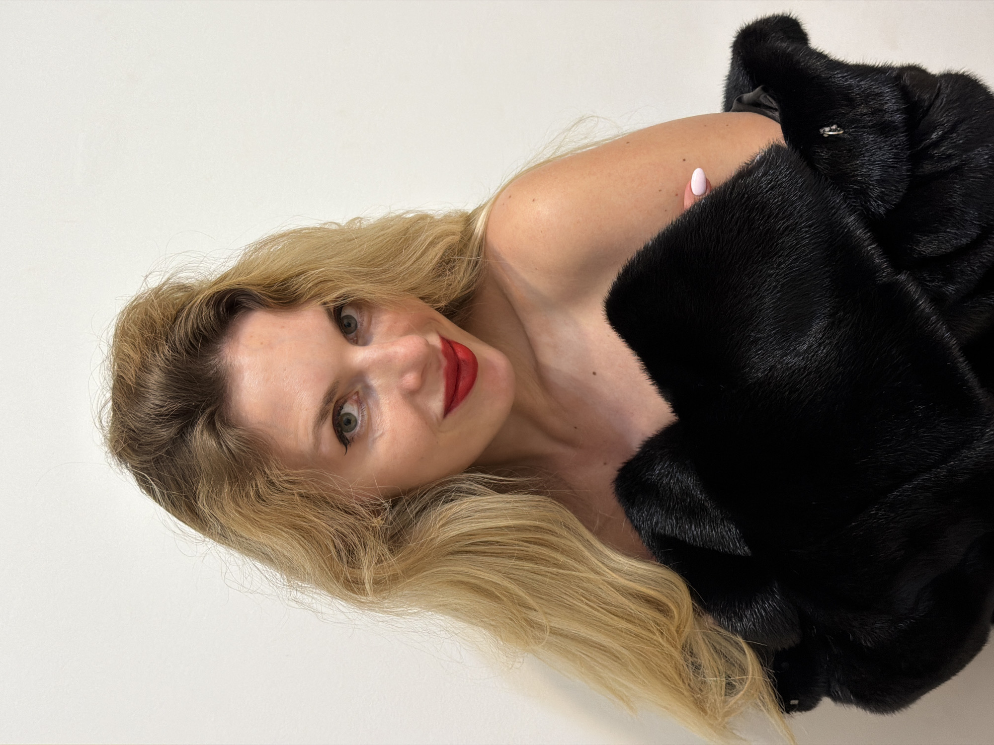 The image shows a woman with blonde hair and red lipstick wearing a black fur coat. The background is a plain white wall. The photo's orientation is tilted. There's no indication of Budva, Montenegro, in the image.