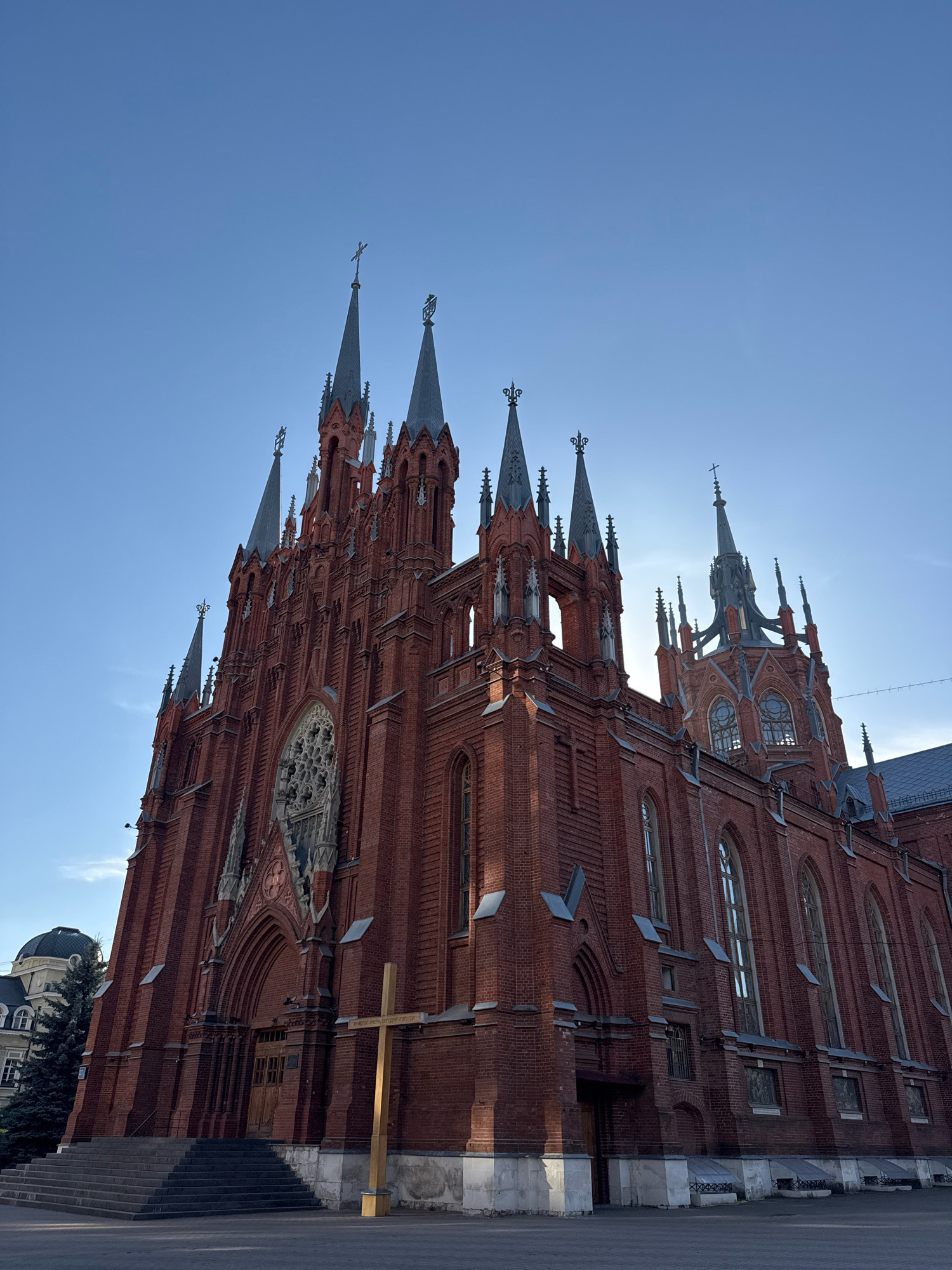 The image shows a low-angle, full shot of the Moscow Cathedral of the Immaculate Conception, a neo-Gothic Roman Catholic cathedral located at Malaya Gruzinskaya Street, Building 22, in the Presnensky District of Moscow, Russia. The cathedral is constructed primarily of red brick and features numerous spires and Gothic architectural elements, including arched windows and a large rose window above the main entrance. A tall, simple wooden cross stands to the right of the entrance. The sky is clear and blue with a slight haze. In the background to the left, a portion of another building with a dark dome is visible, as is a tall evergreen tree. The ground in front of the cathedral is paved with cobblestones. The scene is devoid of any people or animals.