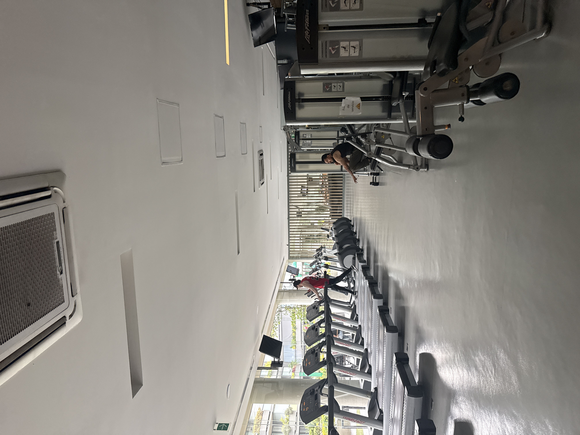 A brightly lit gym in Mexico City, Mexico, features a row of treadmills on the right side with several people using them. The left side of the image shows a white ceiling with rectangular recessed lights and a large air conditioning unit. At the top of the image, gym equipment is visible, including a Life Fitness machine, with a person using it. The overall impression is of a modern, well-equipped gym.