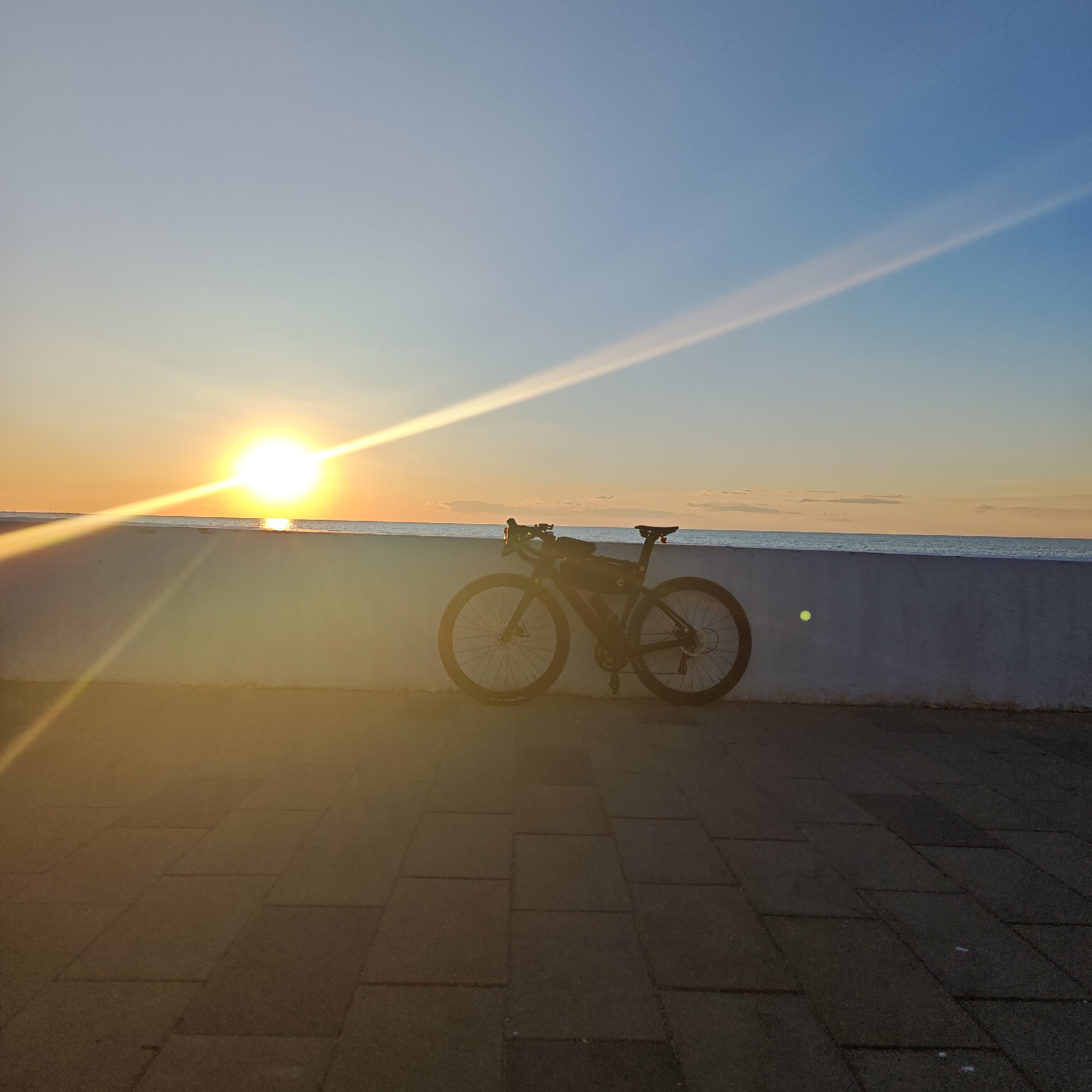 The image depicts a serene coastal or lakeside scene during either sunrise or sunset. A dark-colored bicycle, possibly a road or gravel bike, is the central subject, leaning against a low, light-colored wall that separates a paved promenade from a calm body of water. The bicycle is equipped with frame bags, suggesting it is set up for touring or bikepacking, and features disc brakes.
The sun, a brilliant and intense orb, is positioned low on the horizon, casting powerful lens flares and prominent diagonal rays of light that extend across the sky. The sky transitions from vibrant orange and yellow hues near the sun to a soft, clear blue further away, indicating an absence of heavy cloud cover. The sun's light also reflects brightly on the surface of the water directly beneath it, creating a shimmering effect.
In the foreground, a paved walkway made of large, rectangular paver stones stretches across the bottom of the frame, bathed in the warm, golden light of the sun. The low wall acts as a clear divider between the paved area and the expanse of water. No people are visible, and the scene evokes a sense of peace and solitude. There is no visible text in the image.