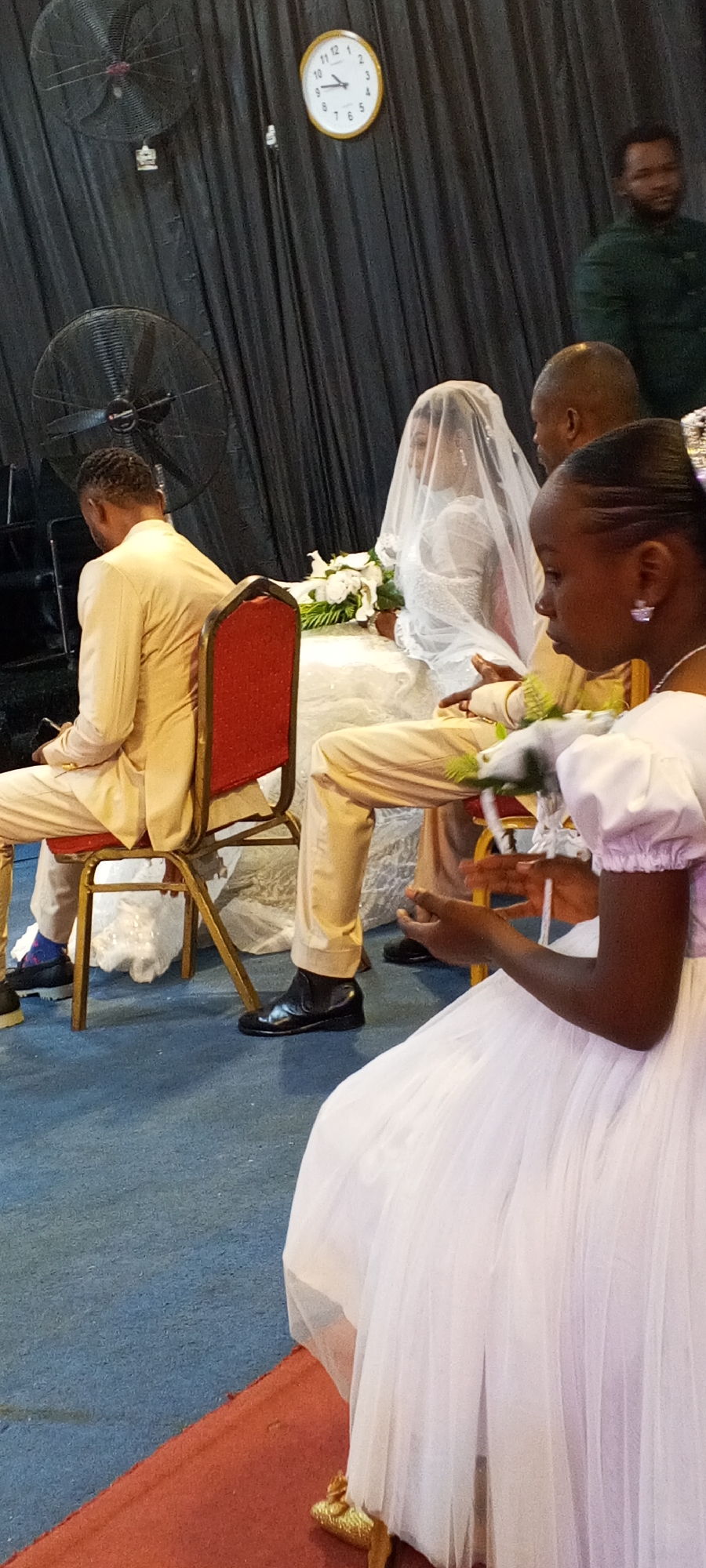 The image appears to be taken indoors, likely at a wedding ceremony in Sabon Tasha, Nigeria, as suggested. A bride wearing a white wedding gown with a veil sits next to a groom in a tan suit. An attendant or bridesmaid in a white dress sits in the foreground. Another man in a tan suit is seen to the left of the bride. A man in green stands behind the bride. The background consists of black curtains, a clock that appears to be displaying 1:08, and ceiling fans. The floor is blue.