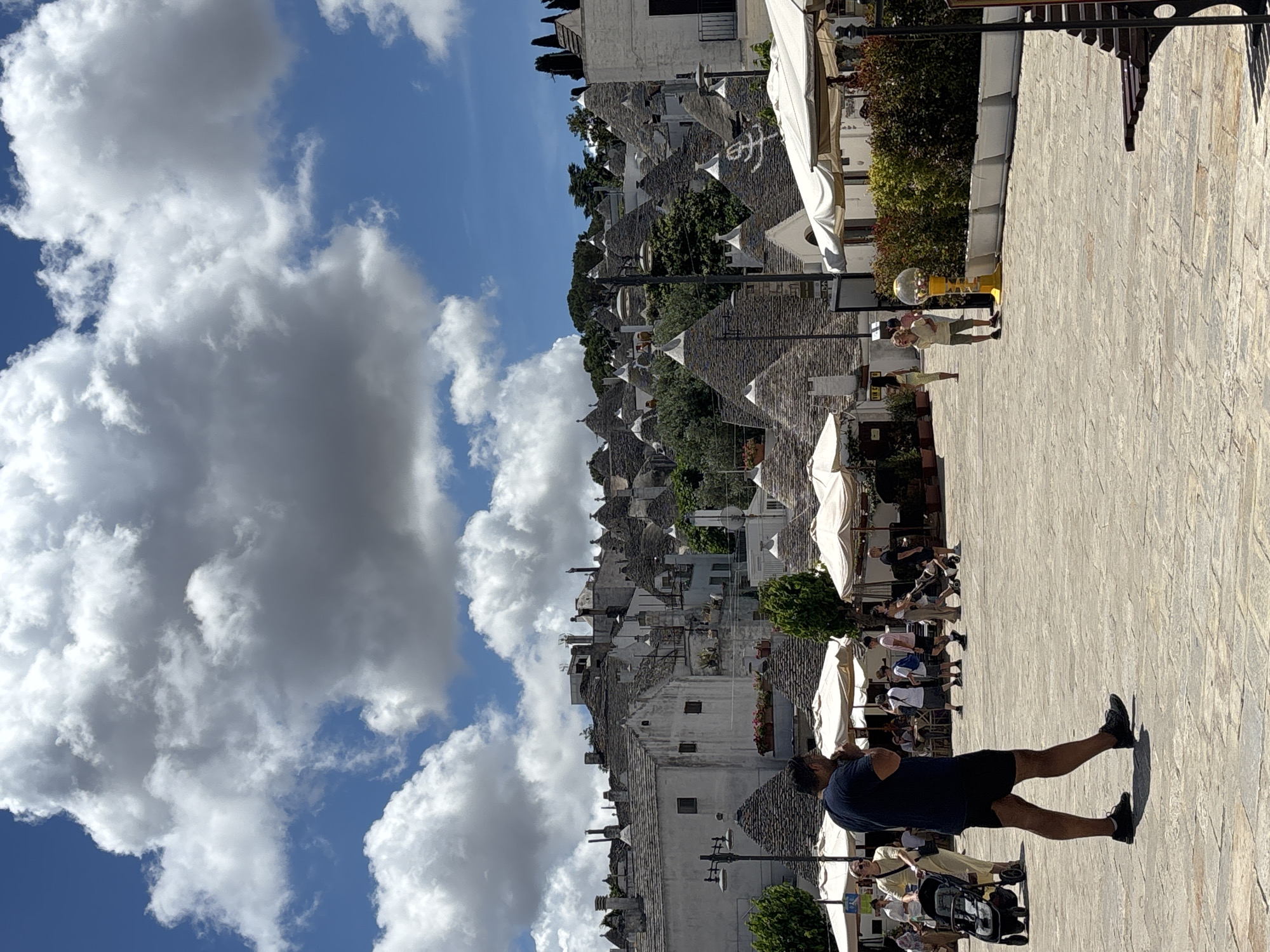 This image captures a vibrant daytime scene in Alberobello, Italy, showcasing its distinctive trulli architecture. The setting is a paved pedestrian area or square, with numerous trulli houses populating the hillside in the background.
The sky is a bright blue, adorned with large, fluffy cumulus clouds that cast varying shades of white and grey. Strong sunlight illuminates the scene, creating prominent shadows on the light-colored, block-paved ground.
In the foreground, a man with dark hair, wearing a dark t-shirt and shorts, stands with his back to the viewer, looking towards the trulli houses and other people. His long shadow stretches behind him. Several other individuals are scattered throughout the mid-ground and background; some are seated at tables under white awnings, indicative of outdoor cafes or restaurants, while others appear to be walking. A woman in a light-colored outfit stands near a bright yellow, spherical vending machine (possibly a gumball machine). Further left, another person is seen pushing a stroller.
The background is dominated by the iconic trulli houses, characterized by their whitewashed walls and unique conical stone roofs, some adorned with symbols. These structures are clustered together, ascending the terrain, with interspersed green trees and bushes providing natural accents. A few traditional buildings with balconies and windows are also visible among the trulli.
The scene suggests a lively atmosphere, likely involving tourists and locals enjoying a sunny day, strolling through the historic town, or relaxing at outdoor establishments. No discernible text is visible in the image.