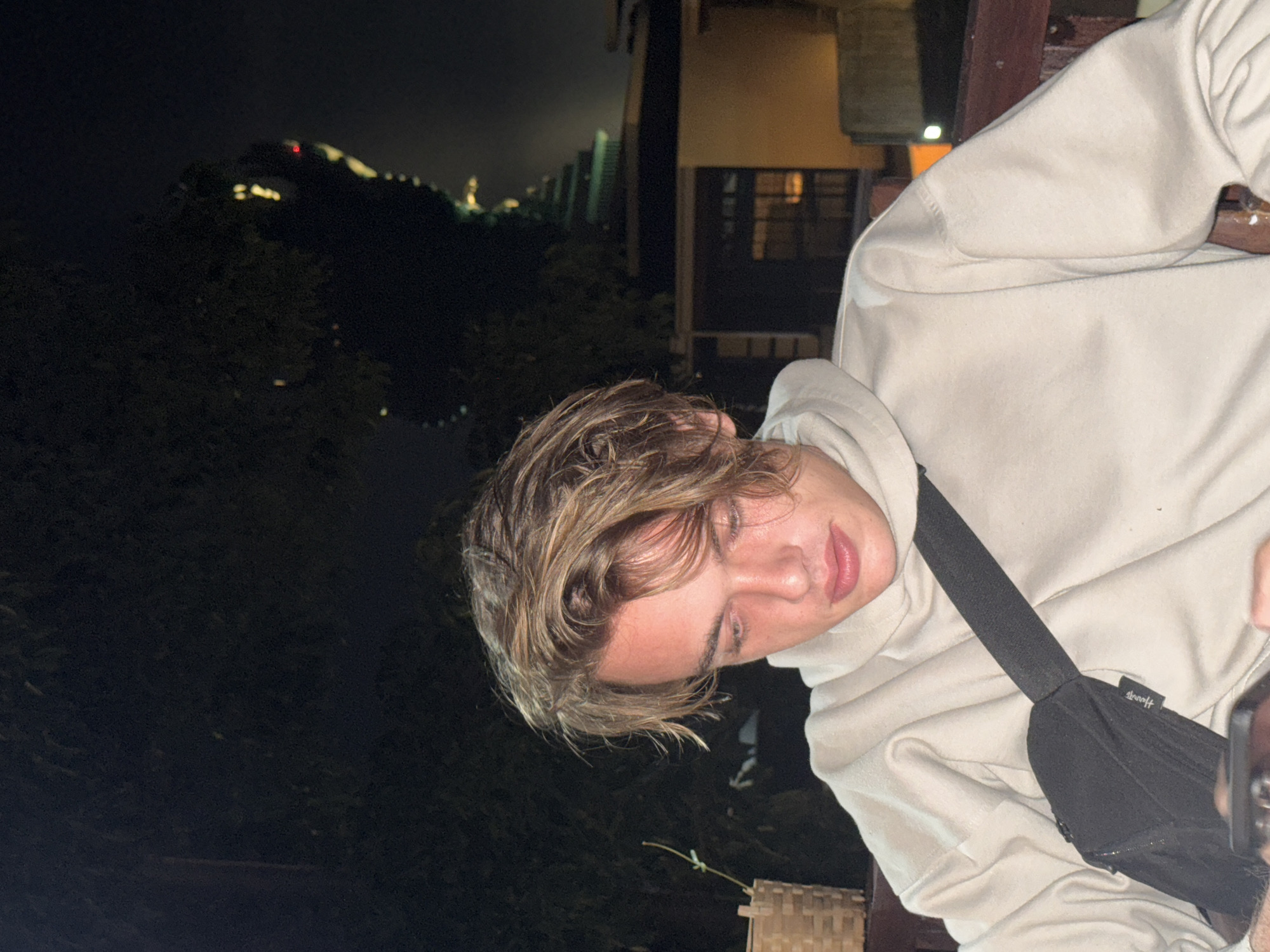 The image, rotated 90 degrees clockwise, captures a young person at night, likely on a balcony or similar elevated outdoor space in Kuta Selatan, Indonesia. The subject has fair skin and light brown, somewhat messy hair, and is wearing a light beige or off-white hooded sweatshirt. Their eyes are cast downwards and slightly to their right, with a calm, neutral expression.
A black sling bag is worn across their chest, with the word "Harrods" clearly visible in white text on its surface. A dark object, possibly a smartphone, is partially visible in the bottom right corner, seemingly being held by an unseen hand. A wooden beam or railing frames the upper right portion of the image, and a piece of woven furniture is visible in the bottom left.
The background is dominated by the dark night sky. Distant city lights or illuminated structures are scattered across a hilly landscape, with a notable red light prominent on one of the hills. Closer to the foreground, dark trees and foliage are discernible. To the immediate right, part of a building with a dimly lit window adds to the setting. The scene depicts a casual, nocturnal moment.