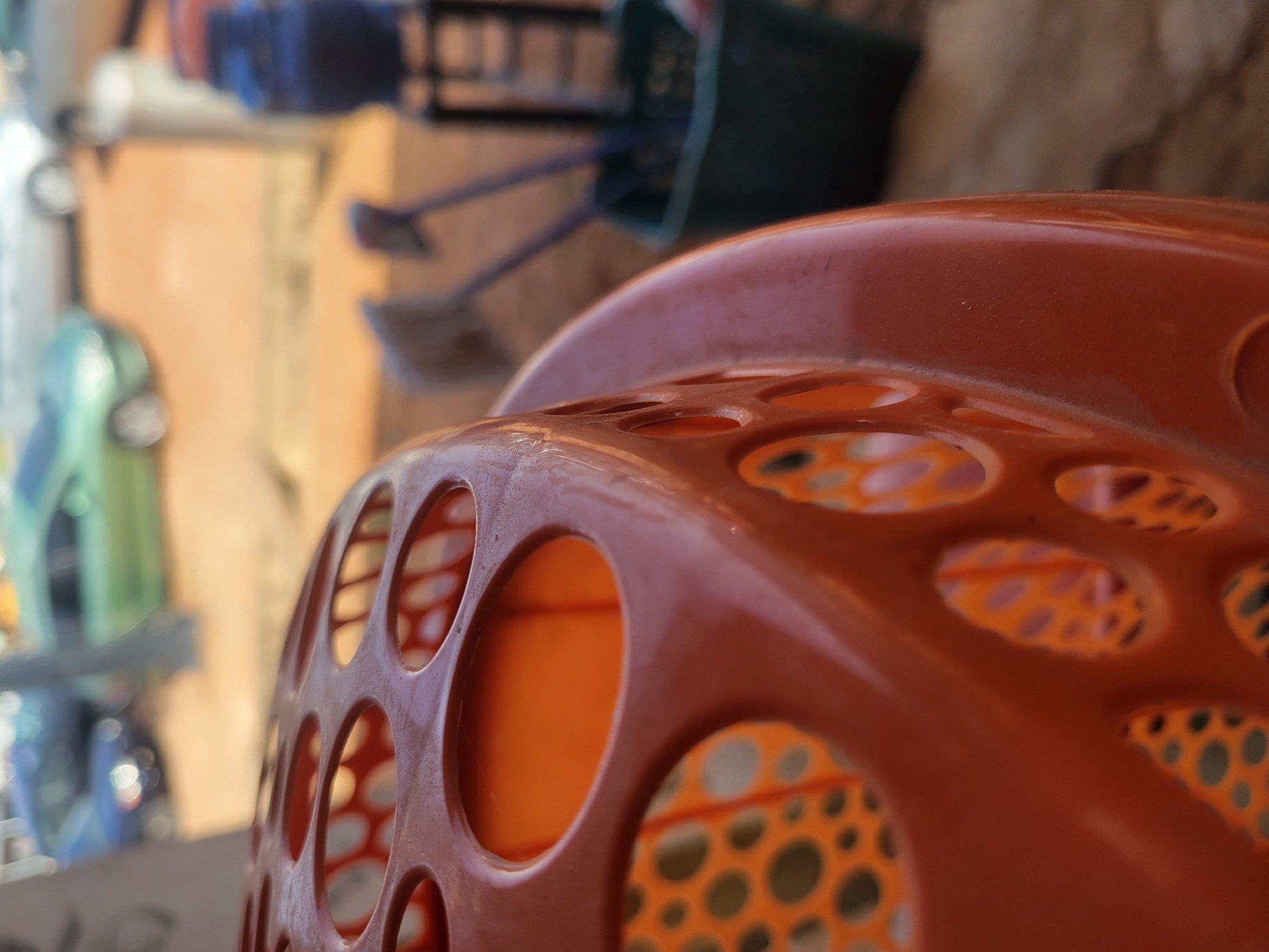 The image features a close-up view of an orange, round, plastic basket or container with numerous circular holes. The container is the primary focus of the image. The background is blurred, but suggests an outdoor or semi-outdoor environment. Based on the additional data, there appear to be other items, including containers and tools, in the blurred background. The specific city (Jos, Nigeria) does not have a visible effect on the image's content.