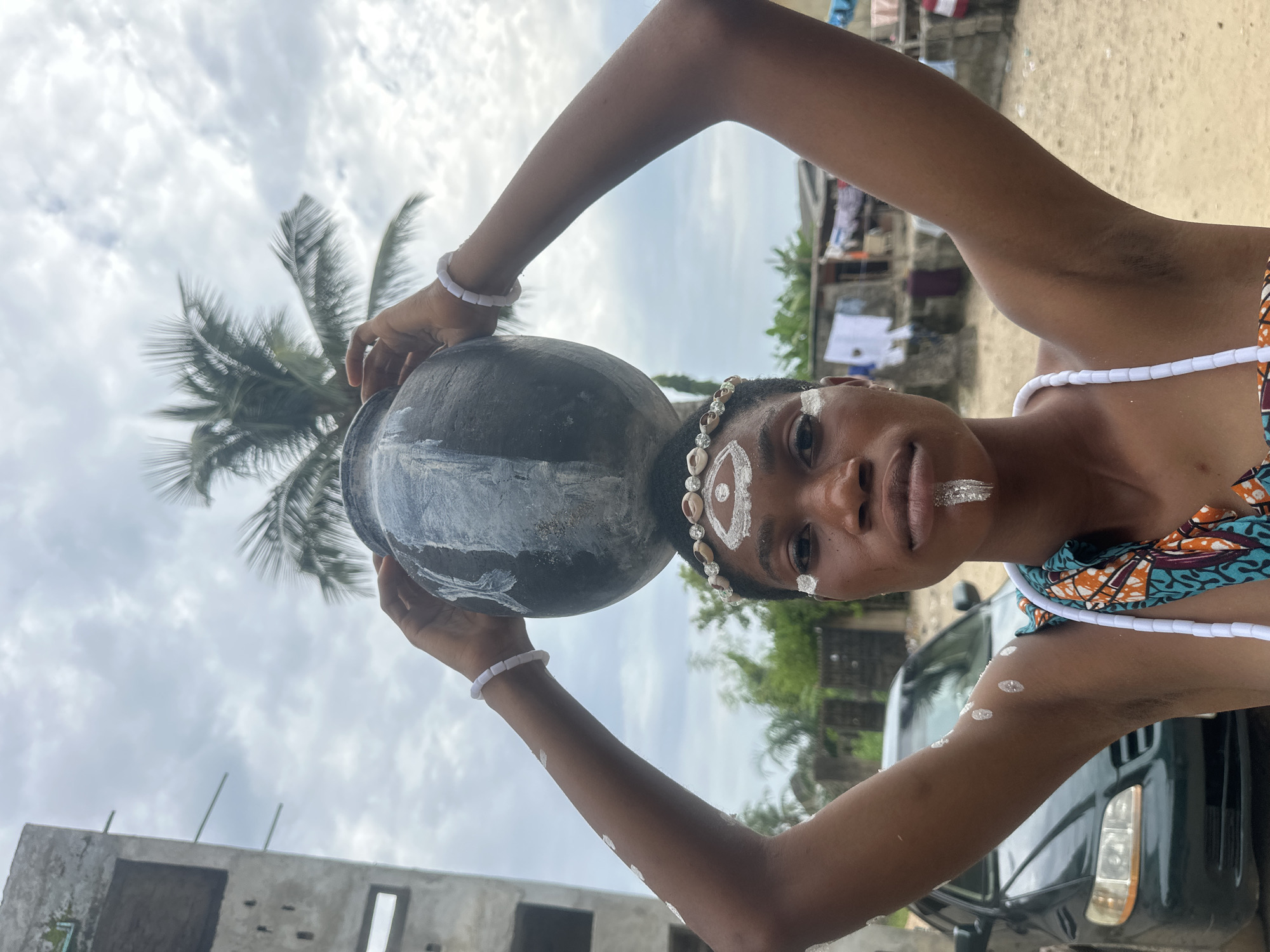 A young Black woman stands outdoors, balancing a dark clay pot on her head. She is adorned with white body paint, including markings on her forehead, under her eyes, and on her chin, resembling a stylized eye and a beard. She wears a necklace of white beads and a headband decorated with shells. Her arms are raised, holding the pot steady. The background shows a partially constructed building to the left, a dark green car parked to the left, and several other structures, some with laundry hanging outside, suggesting a residential area. A palm tree is visible behind the pot. The sky is cloudy. The scene appears to be set in the city of Badagry, Nigeria, based on contextual information.