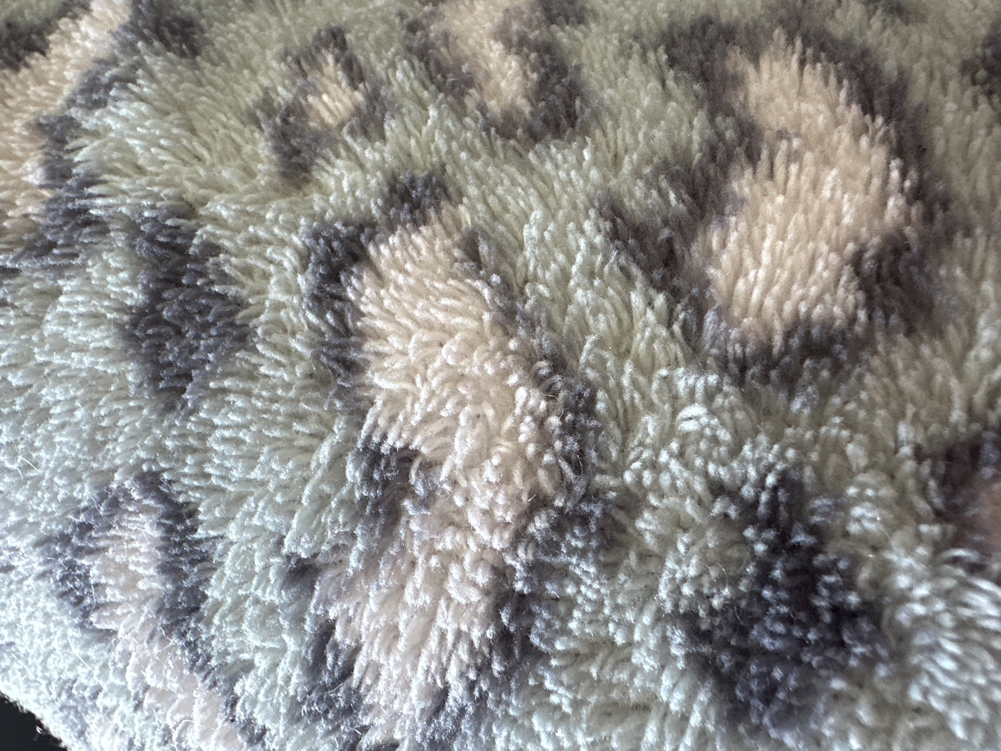 The image displays a close-up, top-down view of a deep-pile textile surface. The fabric presents a mottled, irregular pattern comprising shades of off-white, light tan/beige, and dark gray/charcoal. Individual fibers, appearing shaggy, soft, and varying in height, create a dense, textured surface. The darker gray elements form irregular, amorphous shapes and splotches against the lighter background hues. Direct, directional lighting illuminates the upper portions of the fibers, producing subtle highlights and shadows that emphasize the texture. No individuals, distinct objects beyond the textile, or specific actions are discernible within the frame. The location within Huixquilucan de Degollado, Mexico, cannot be determined from the visual information provided in the image.