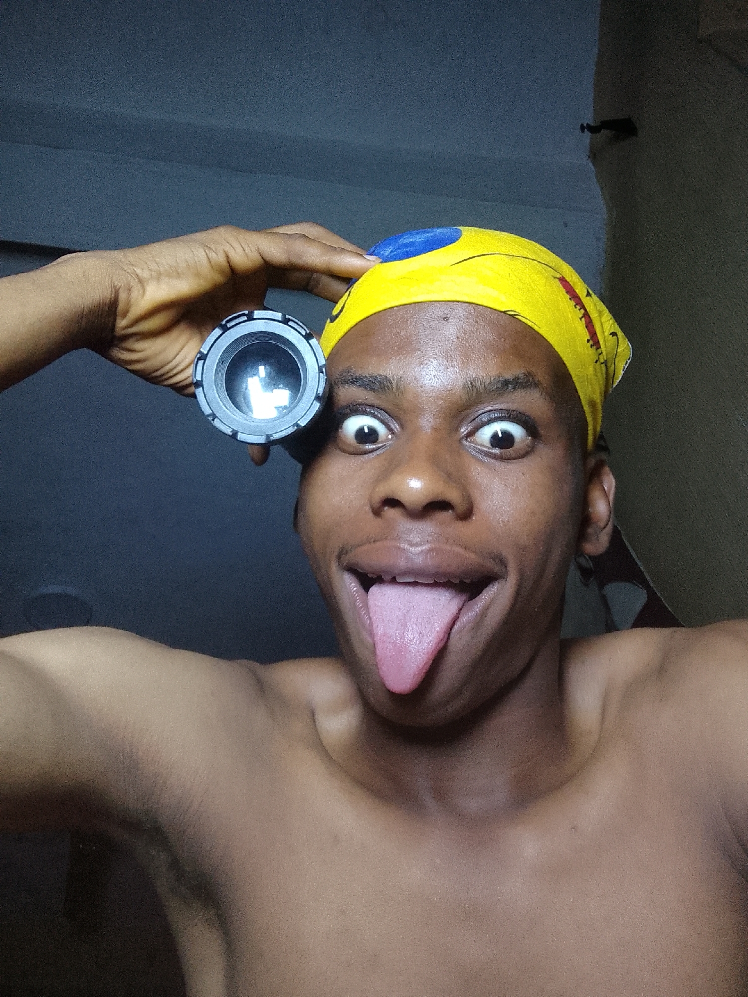 A shirtless, dark-skinned individual with a yellow and blue bandana is looking directly at the camera with wide eyes and their tongue extended. They are holding a cylindrical object with a lens over their head. The background is a dimly lit wall. Based on the request for the location to be Nkpor, Nigeria, the scene is likely occurring in a residential or personal space within Nkpor, Nigeria.