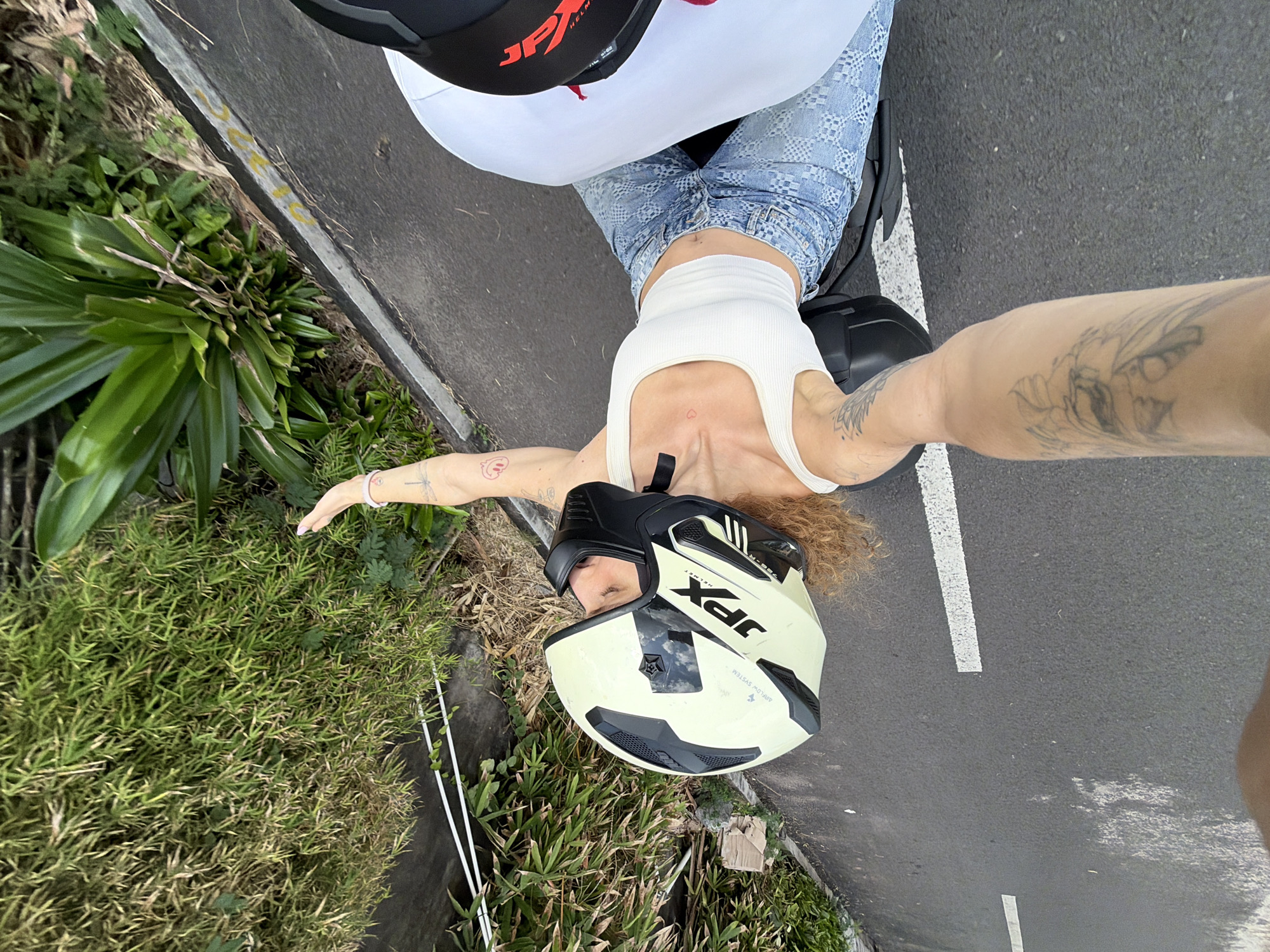 A woman is riding a motorcycle on a paved road, possibly in Kediri, Indonesia, given the presence of lush greenery and bamboo plants lining the roadside, indicative of a tropical or subtropical climate. The road features white lane markings. The woman is wearing a cream-colored sleeveless top, blue jeans with a patterned design, and a white and black motorcycle helmet. She has curly blonde hair visible beneath the helmet and tattoos on her left arm and right forearm. Her right arm is extended outwards. The scene is captured from a high-angle perspective, suggesting the photo was taken by someone on the same motorcycle.