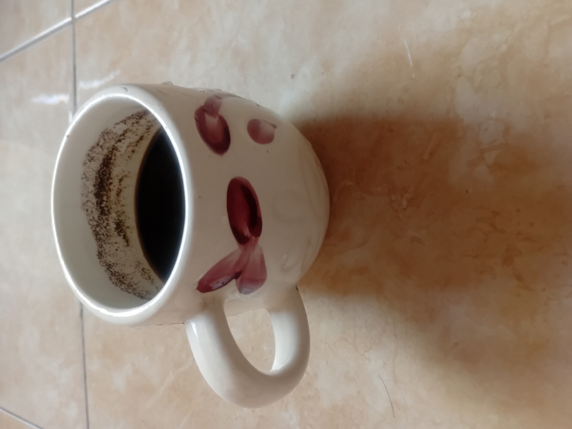 A close-up, eye-level shot shows a white coffee mug with a handle resting on a tiled floor. The mug contains dark liquid coffee with some coffee grounds visible along the rim of the interior. The mug is decorated with red petal-like designs. The tiled floor is a light brown color with visible grout lines. The lighting suggests an indoor setting, possibly a kitchen or dining area. There is a shadow cast by the mug onto the floor.