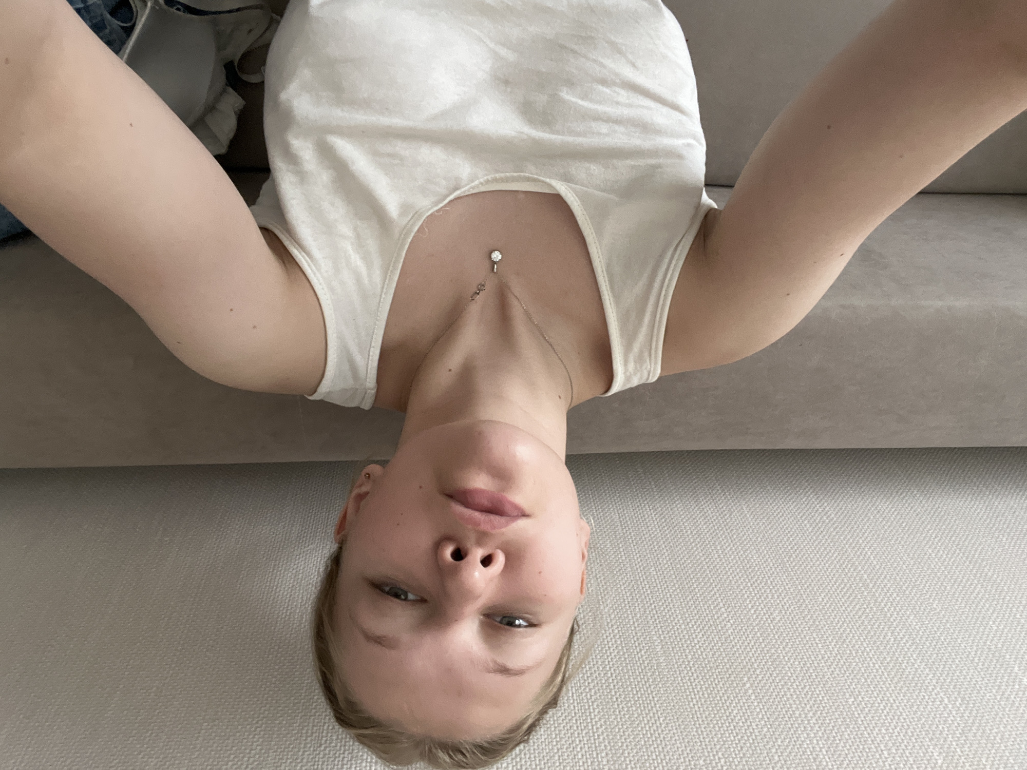 The image is an upside-down selfie taken by a young person with blonde hair, lying on a couch. The subject is wearing a white tank top and a delicate necklace with a clear pendant. The background shows a textured couch and a wall. The lighting suggests it is daytime, possibly indoors. The subject's expression is neutral. The location context is Perm, Russia. There is no visible text in the image.
