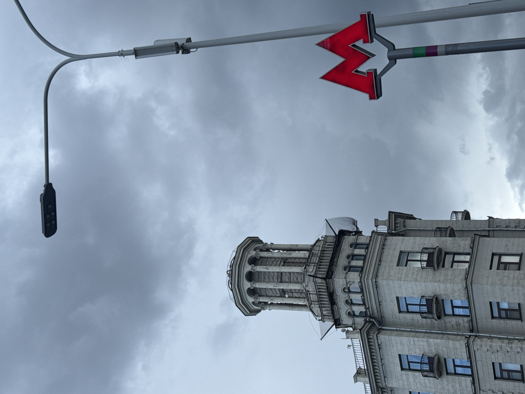 The image shows a view in Moscow, Russia, featuring architectural elements against a cloudy sky. In the foreground, there is a red "M" sign, which indicates the Moscow Metro. To the left of the sign is a modern street lamp. Below these, the upper section of a building with a detailed, turret-like structure and several windows is visible. The cloudy sky provides a muted backdrop to the architectural details.