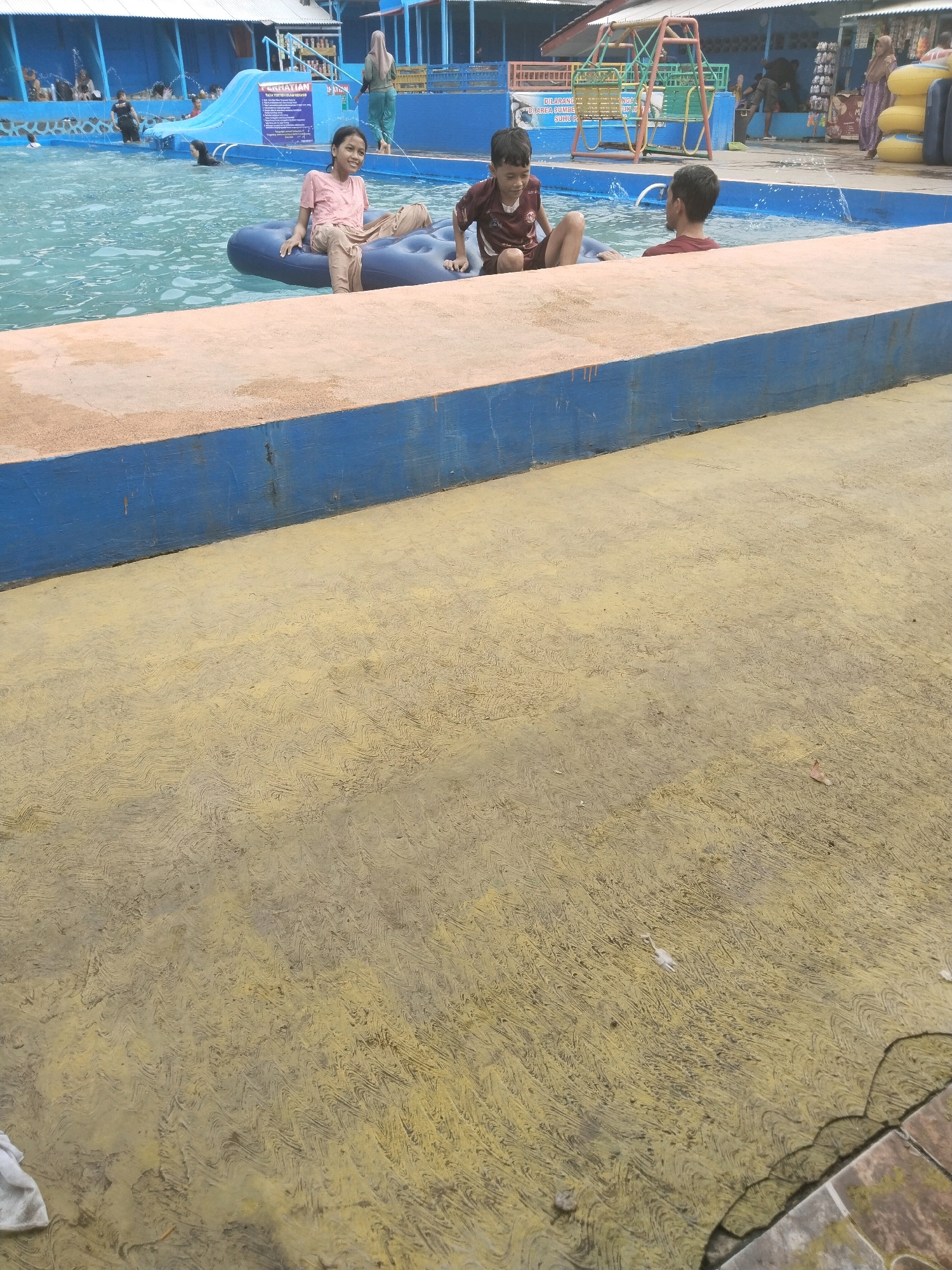 In the city of Batukuwung, Indonesia, a group of three children is seen at a water park. Two boys and a girl are sitting on an inflatable raft in a swimming pool. The girl, on the left, is wearing a pink t-shirt and beige shorts. The boy next to her is wearing a maroon and white t-shirt and maroon shorts. The third boy, partially visible on the right, is wearing a red t-shirt. The pool is surrounded by a concrete deck that transitions to a yellow, textured pavement. In the background, a blue and white structure with a waterslide and several people can be observed. The overall scene depicts a recreational activity at a water park.