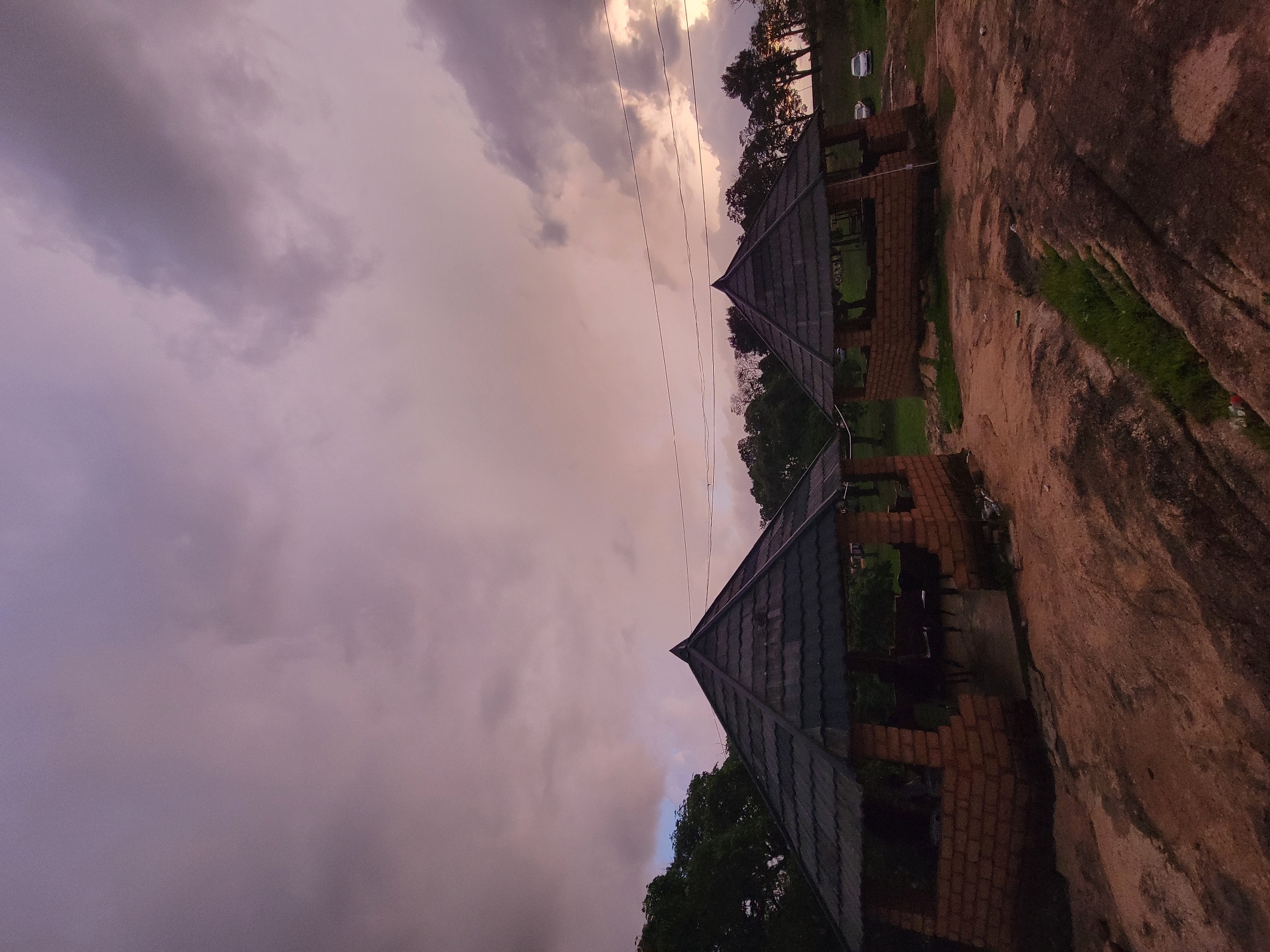 The scene is an outdoor area in an unknown location in the city of Unknown, Unknown, captured under twilight conditions. The sky is predominantly overcast, displaying a gradient of colors from purplish-grey in the upper left to pinkish-orange and golden hues towards the right horizon. Two open-sided, pyramidal-roofed structures, constructed with reddish-brown brick bases and dark tiled roofs, are prominent. The larger structure is positioned on the left, with indistinct dark forms visible within its interior, possibly indicating seated individuals or furniture. A second, smaller structure stands to the right. The foreground and midground consist of a broad expanse of reddish-brown rocky terrain, featuring crevices and scattered patches of green grass. A line of dense trees forms the background, particularly on the right side, where two light-colored vehicles are discernible on a raised area. Overhead electrical wires are visible spanning the upper mid-ground of the image. No specific actions or interactions are clearly discernible.