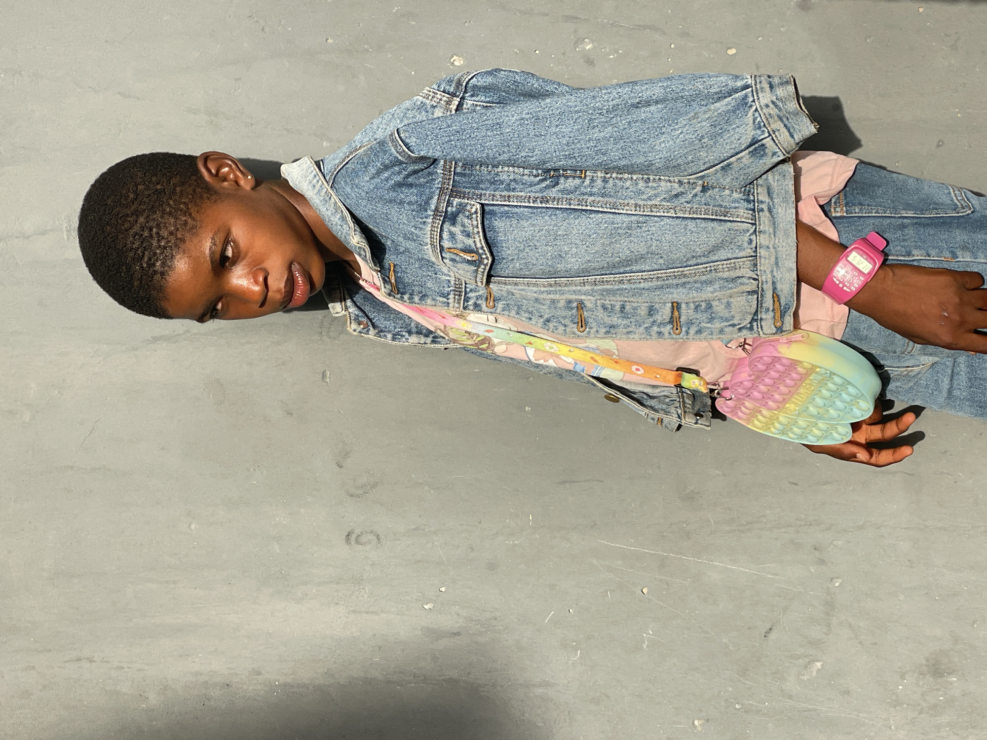 A young individual is captured in a close-up, horizontal shot, lying on a plain, light grey concrete surface in Badagry, Nigeria. The person has short, dark hair and a dark skin tone, with their eyes slightly closed or gazing downwards, conveying a relaxed and serene posture. They are dressed in a light blue denim jacket worn over a light pink t-shirt, with denim jeans visible on the lower right of the frame.
Notable accessories include a vibrant pink digital watch on the right wrist, uniquely designed to resemble an old-fashioned mobile phone, which displays "5:22" on its screen. A small, pastel rainbow-colored bag, styled like a heart-shaped pop-it fidget toy, is slung across their body with a light-colored strap. The text "LOVES YOU" is faintly visible on the bag's strap.
Strong, directional lighting from the upper right casts soft shadows, indicating a bright environment, possibly outdoors or under bright artificial light. The background is a minimalist, unadorned concrete floor with subtle scuff marks and textures, highlighting the subject and their colorful accessories. The overall scene is casual and still.